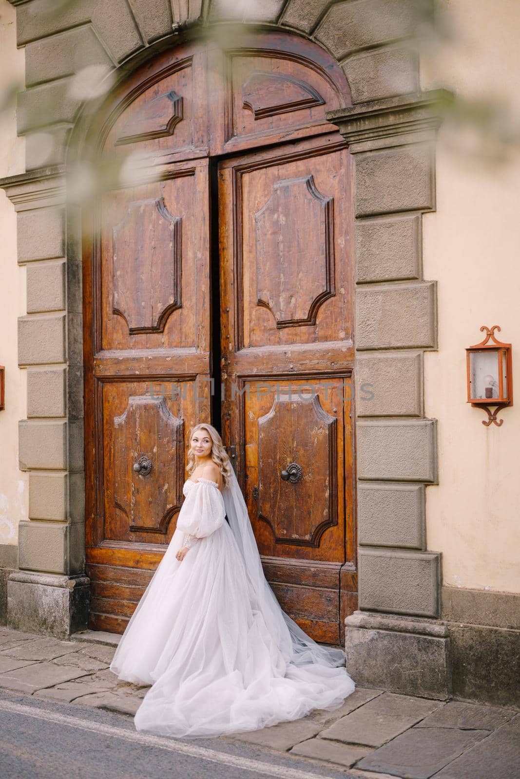 Beautiful bride in a big white wedding dress and long veil is standing in front of huge wooden vintage doors of an old Italian villain Tuscany, Florence. by Nadtochiy