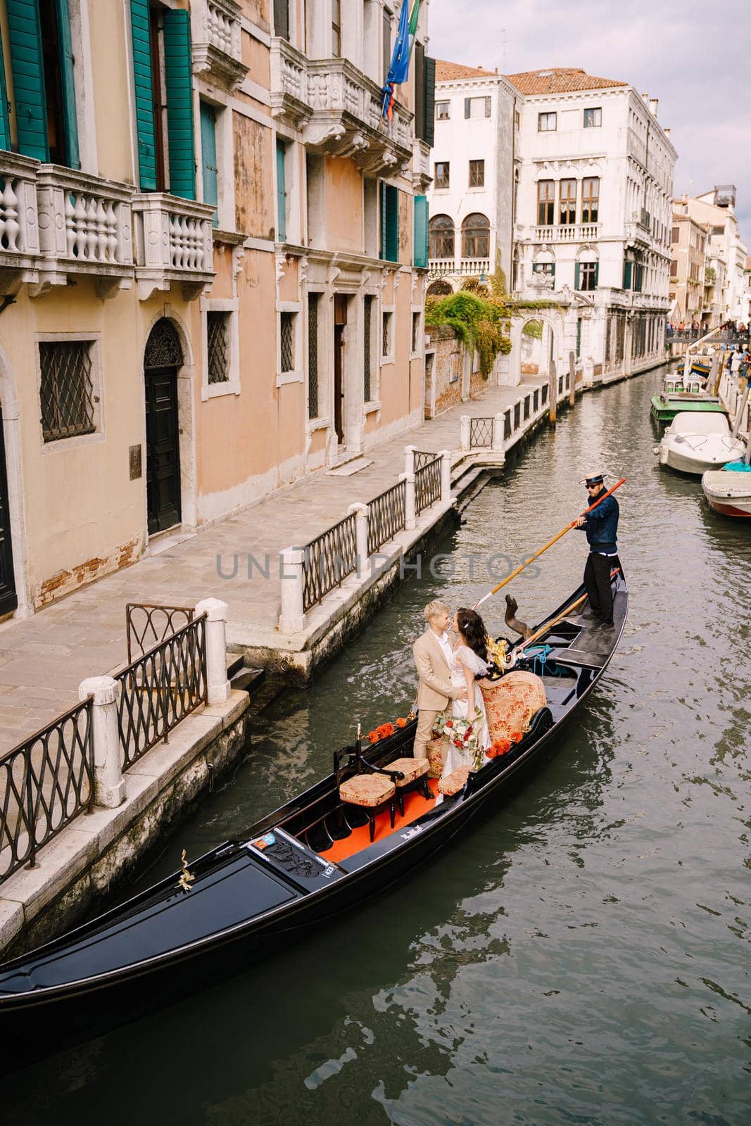 The gondolier rides the bride and groom in a classic wooden gondola along a narrow Venetian canal. Newlyweds standing in a boat against the background of ancient buildings.