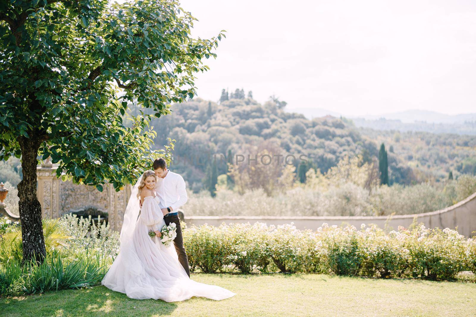 Wedding in Florence, Italy, in an old villa-winery. Bride and groom in the shade of a tree. Wedding couple walks in the garden. by Nadtochiy