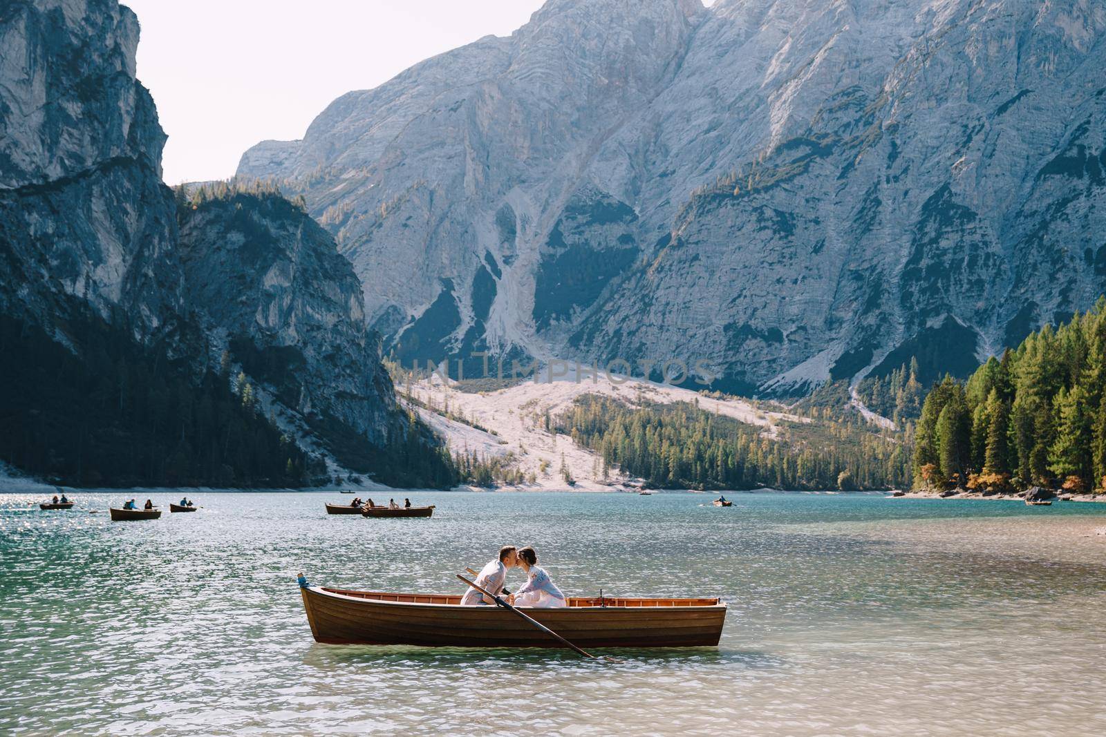 Bride and groom sail in a wooden boat at the Lago di Braies in Italy. Wedding in Europe, on Braies lake. The newlyweds are sitting in the boat and kissing. by Nadtochiy
