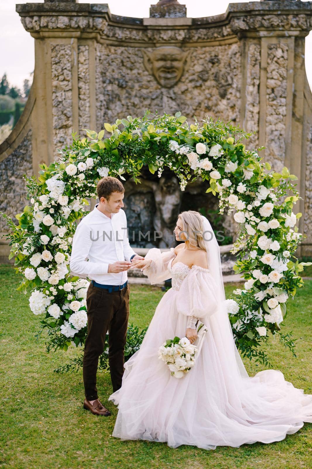 The groom puts the ring on the brides finger. Wedding at an old winery villa in Tuscany, Italy. Round wedding arch decorated with white flowers and greenery in front of an ancient Italian architecture by Nadtochiy