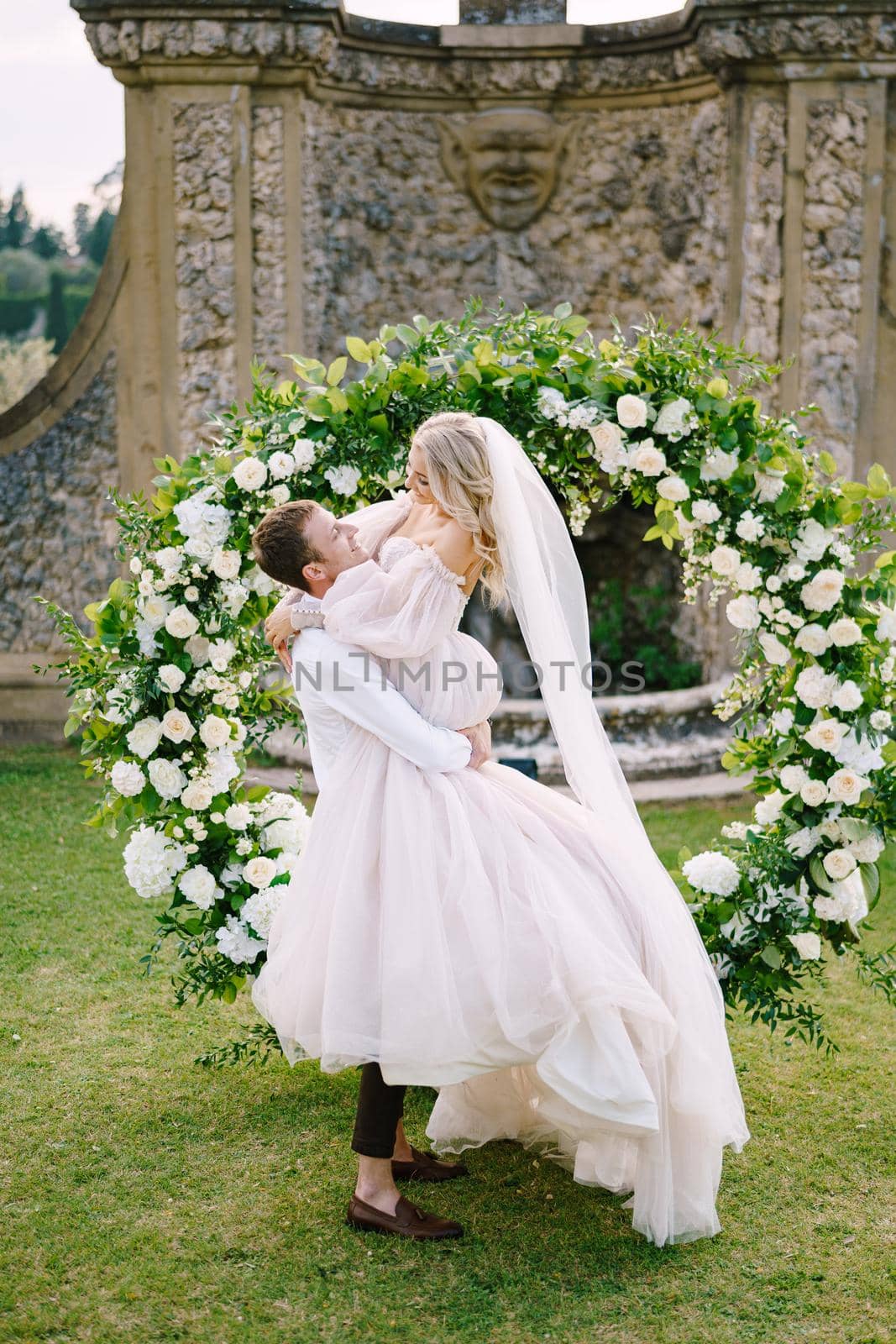 Wedding at an old winery villa in Tuscany, Italy. The groom circles the bride in his arms. Round wedding arch decorated with white flowers and greenery in front of an ancient Italian architecture. by Nadtochiy