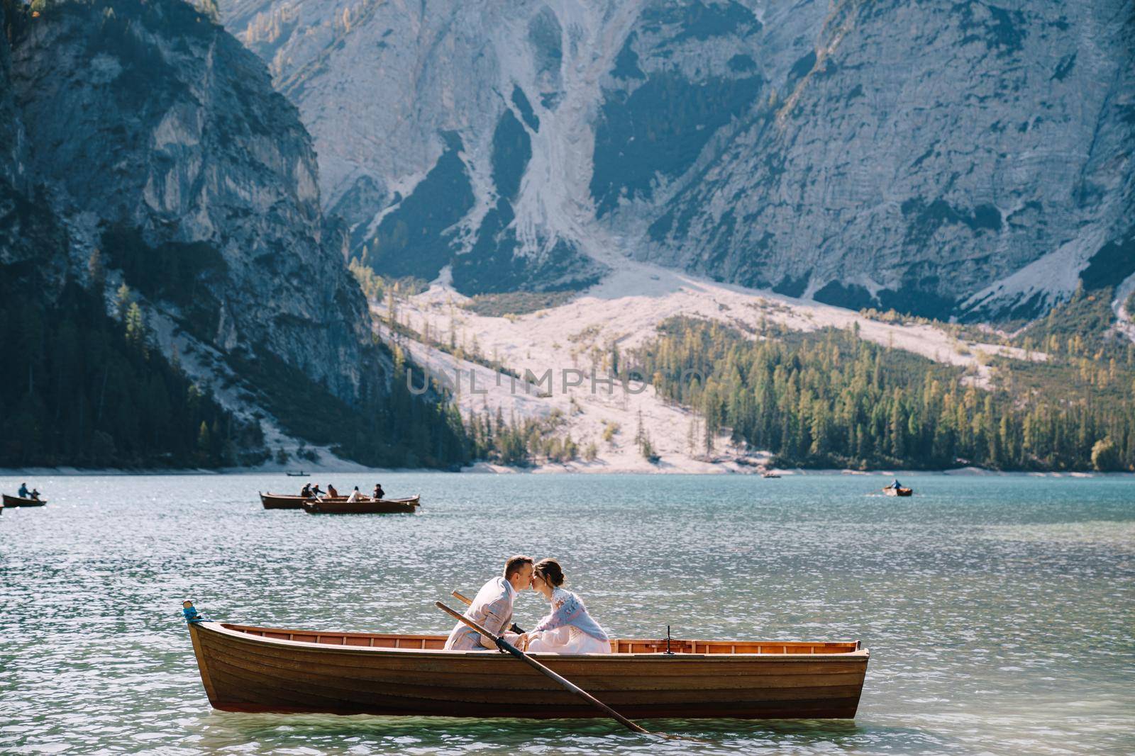 Bride and groom sail in a wooden boat at the Lago di Braies in Italy. Wedding in Europe, on Braies lake. The newlyweds are sitting in the boat and kissing. by Nadtochiy