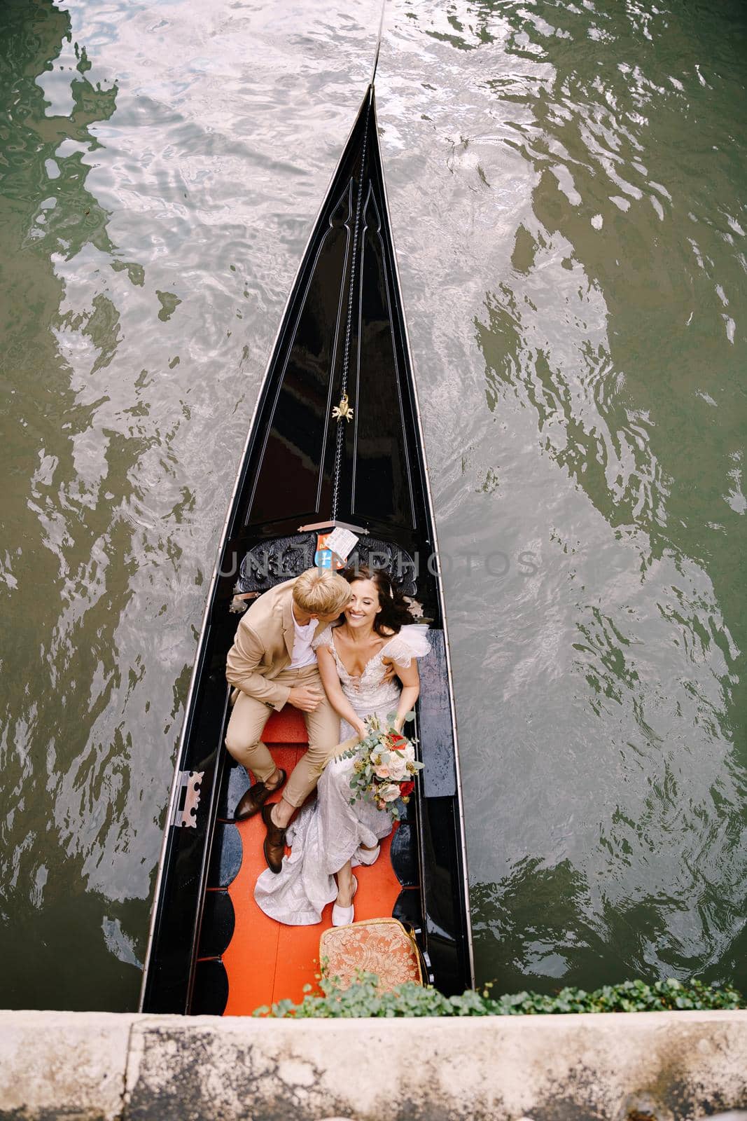 Italy wedding in Venice. A gondolier rolls a bride and groom in a classic wooden gondola along a narrow Venetian canal. Newlyweds are sitting in a boat on the background of ancient buildings. by Nadtochiy
