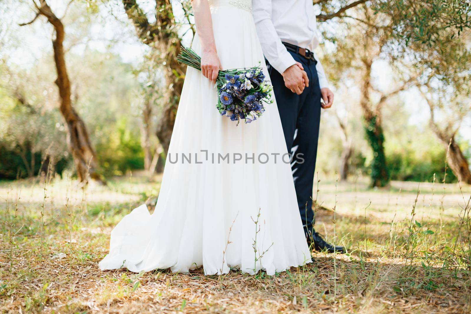 Bride with a bouquet of blue flowers and the groom stand side by side in the olive grove and hold hands by Nadtochiy