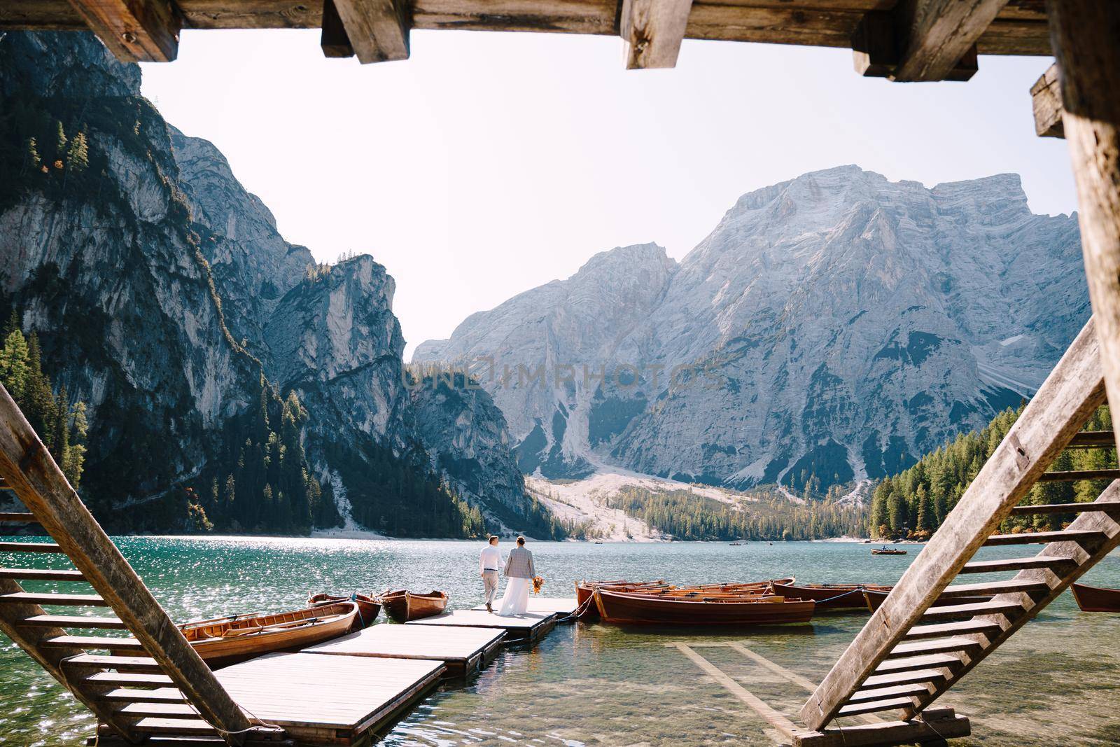 The bride and groom walk along a wooden boat dock at the Lago di Braies in Italy. Wedding in Europe, on Braies lake. Newlyweds walk, kiss, hug on a background of rocky mountains. by Nadtochiy