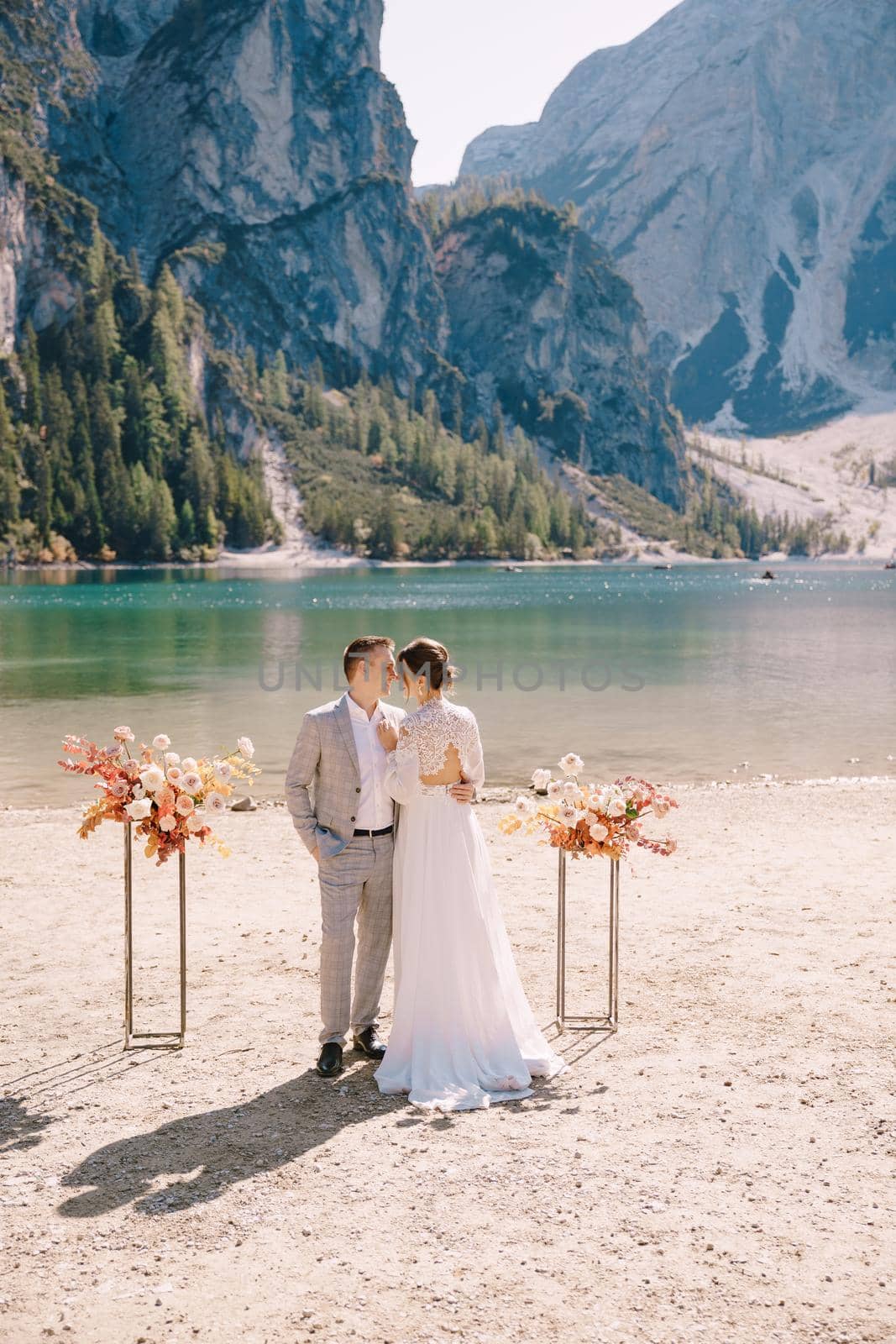 The bride and groom stand still for the ceremony, with an arch of autumn floral columns, against the backdrop of Lago di Braies in Italy. Destination wedding in Europe, at Braies lake.