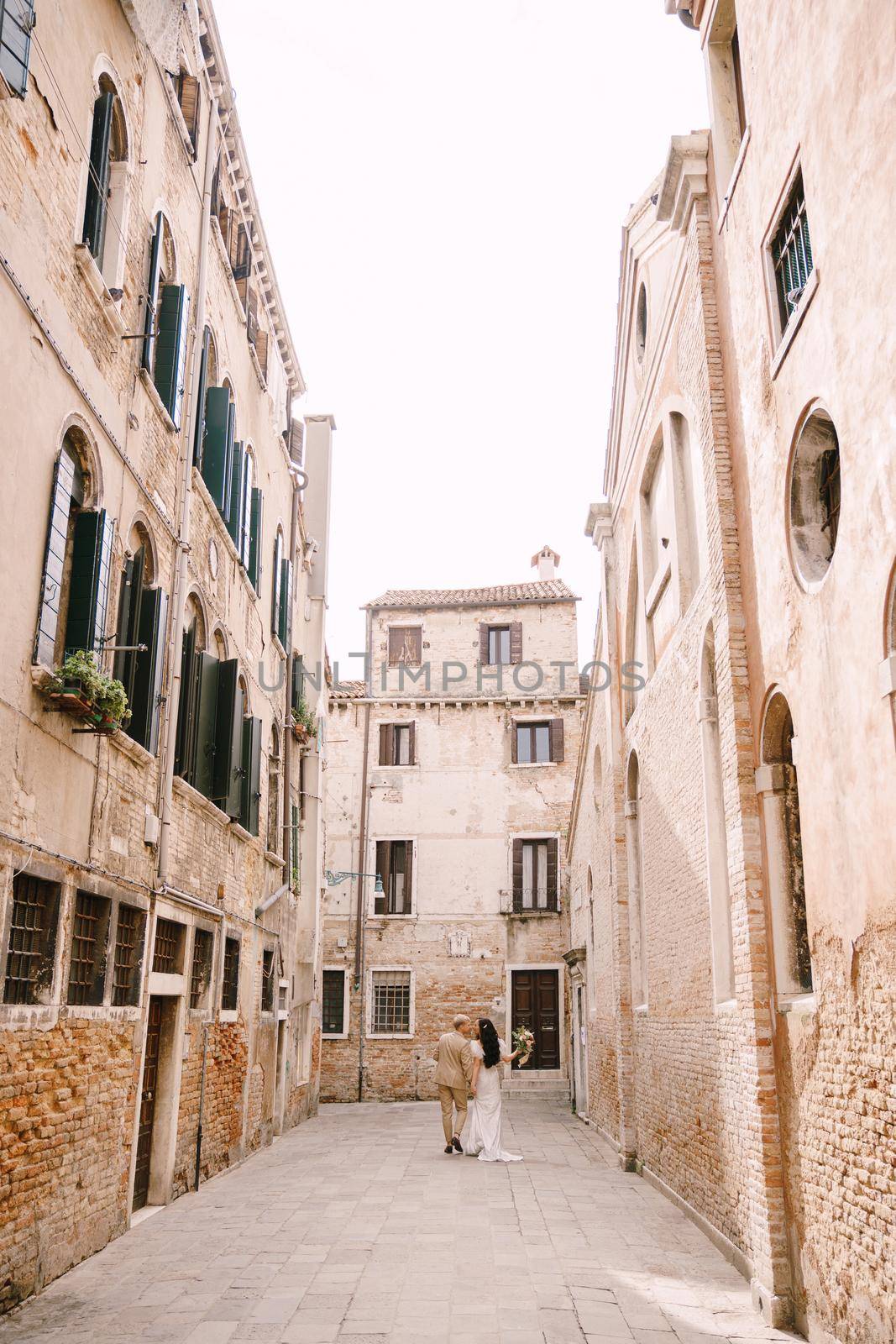 Italy wedding in Venice. The bride and groom walk along the deserted streets of the city. Newlyweds are walking in a dead end alley on the background of brick buildings. by Nadtochiy