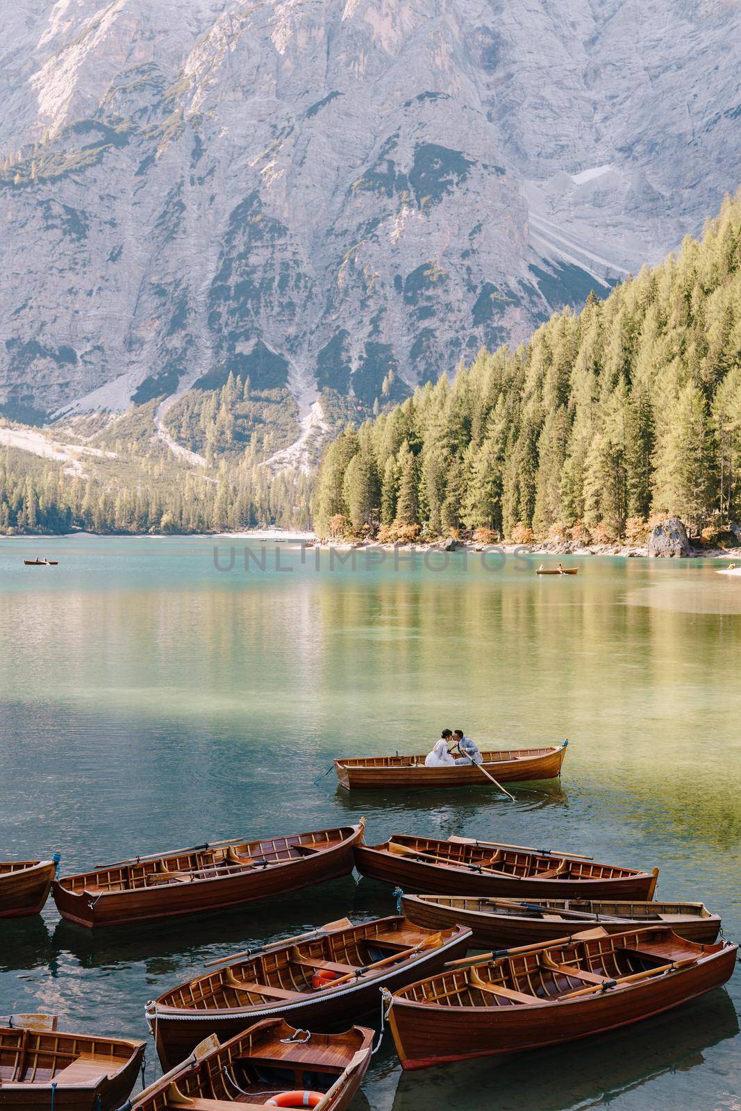 Wedding couple sailing in a wooden boat at the Lago di Braies in Italy. Newlyweds in Europe, on Braies lake, in the Dolomites. The groom rows the oars, the bride sits opposite him. by Nadtochiy