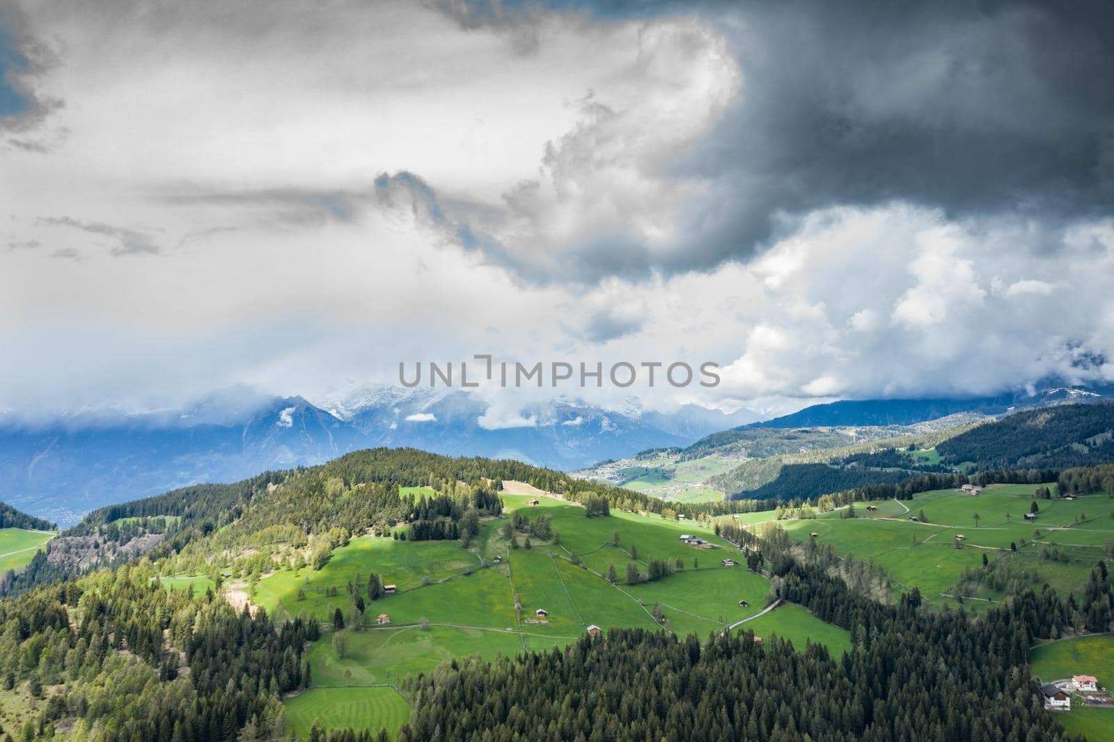 Aerial view of improbable green meadows of the Italian Alps, green slopes of the mountains, Bolzano, huge clouds over a valley, roof tops of houses, Dolomites on background, sunshines through clouds