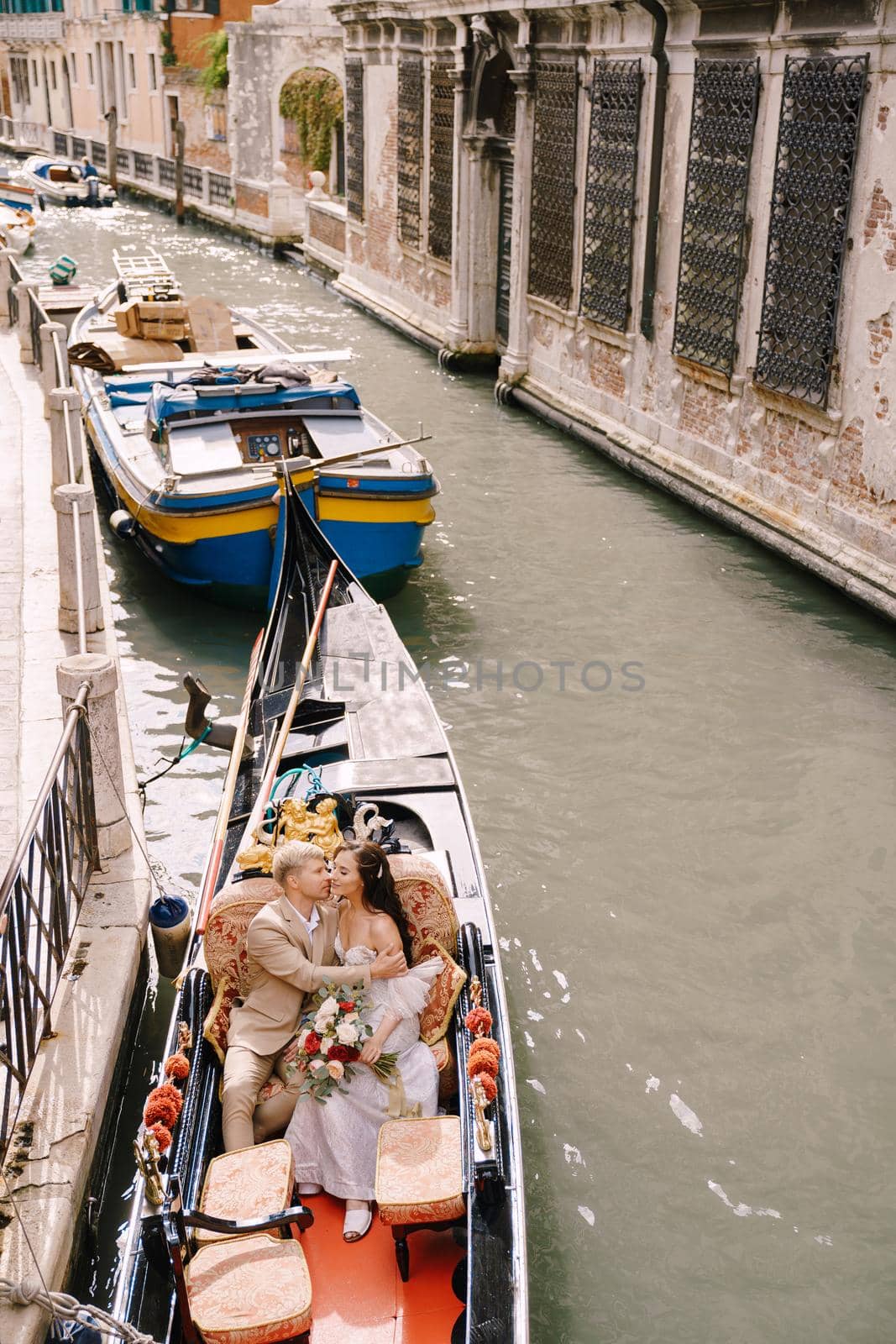 The gondolier rides the bride and groom in a classic wooden gondola along a narrow Venetian canal. Newlyweds kiss in a boat moored to the shore.