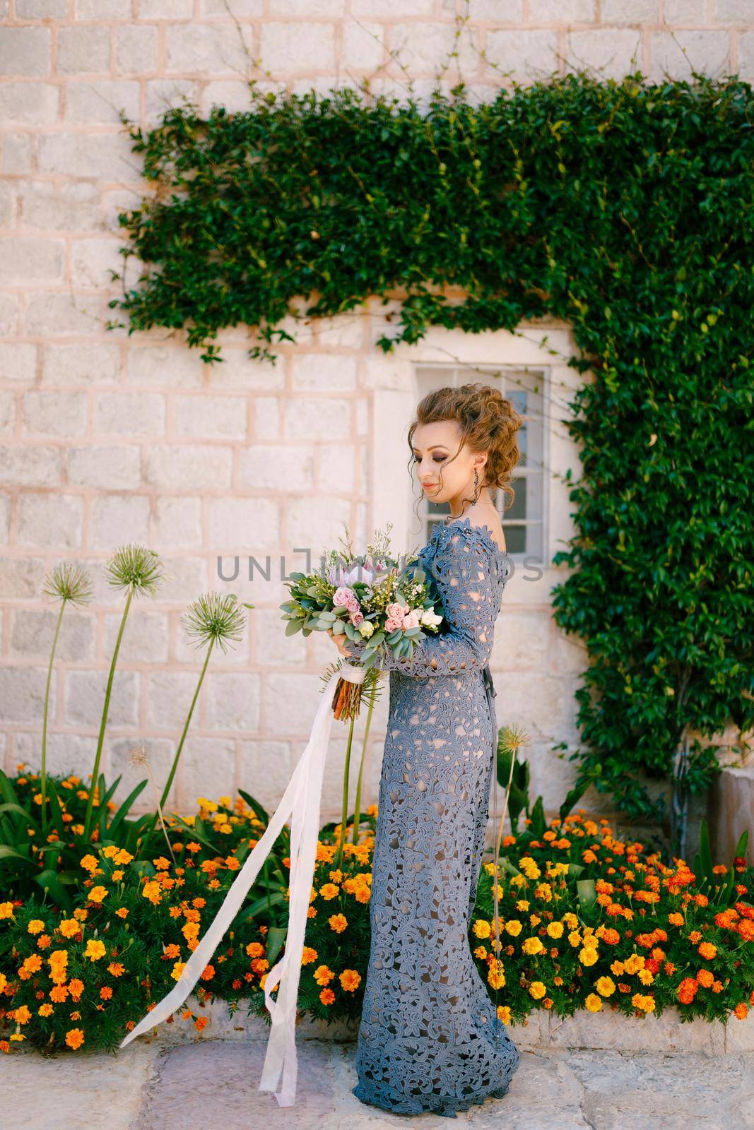 A bride in a blue dress with a bouquet stands at the wall of a house with a green liana and orange flowers, close-up. High quality photo