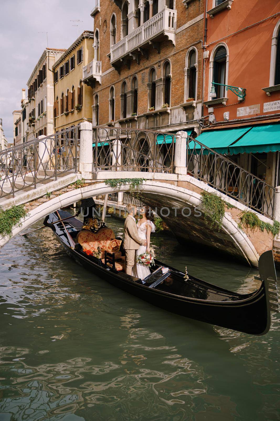 Italy wedding in Venice. A gondolier rolls a bride and groom in a classic wooden gondola along a narrow Venetian canal. Newlyweds in a gondola swim out from under the bridge, stand in a boat and hug. by Nadtochiy