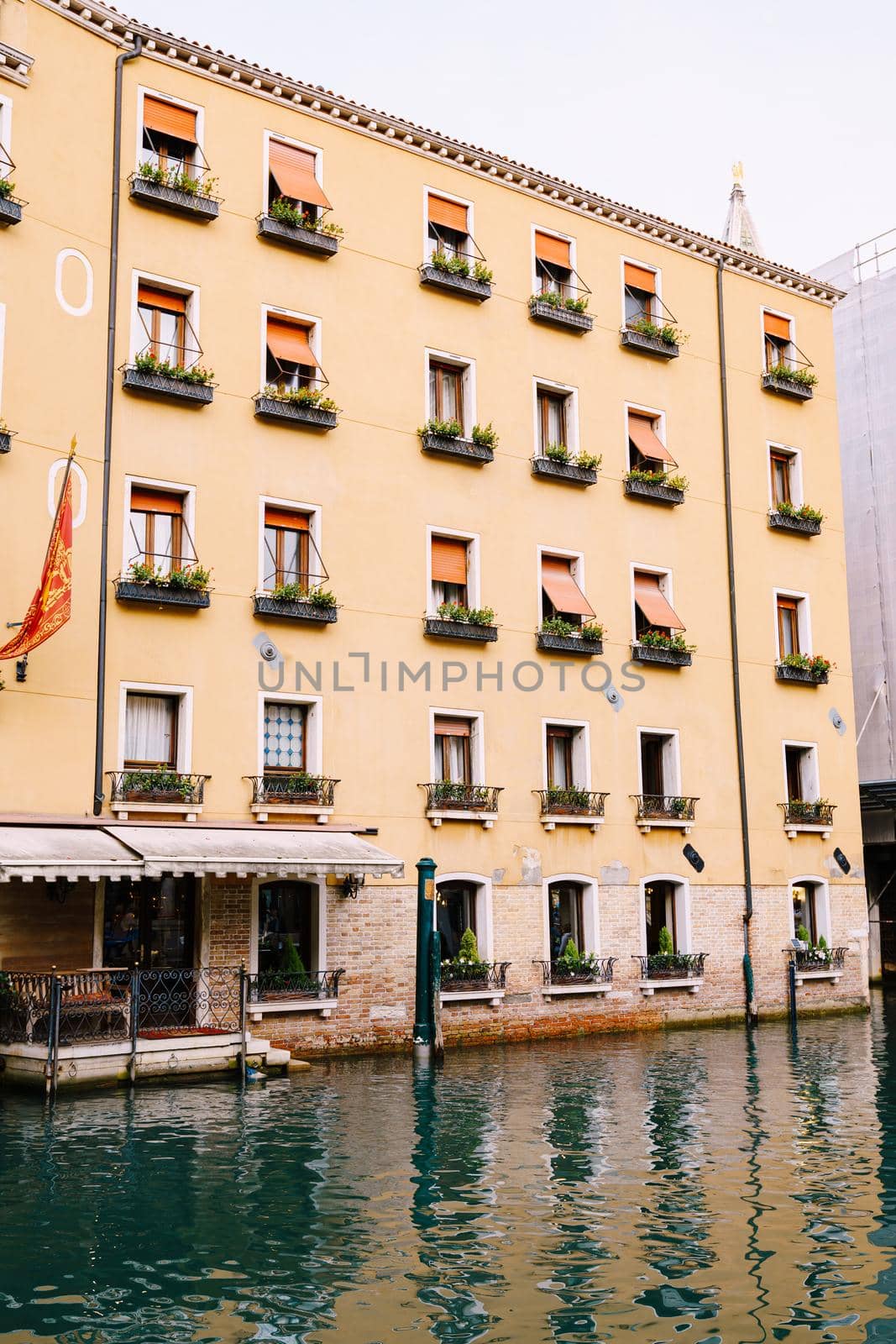 Yellow five-story building in Venice, Italy. The main entrance of the building goes into the water, at entrance there is a sofa, on horseback there are flower pots and orange shutters from the sun