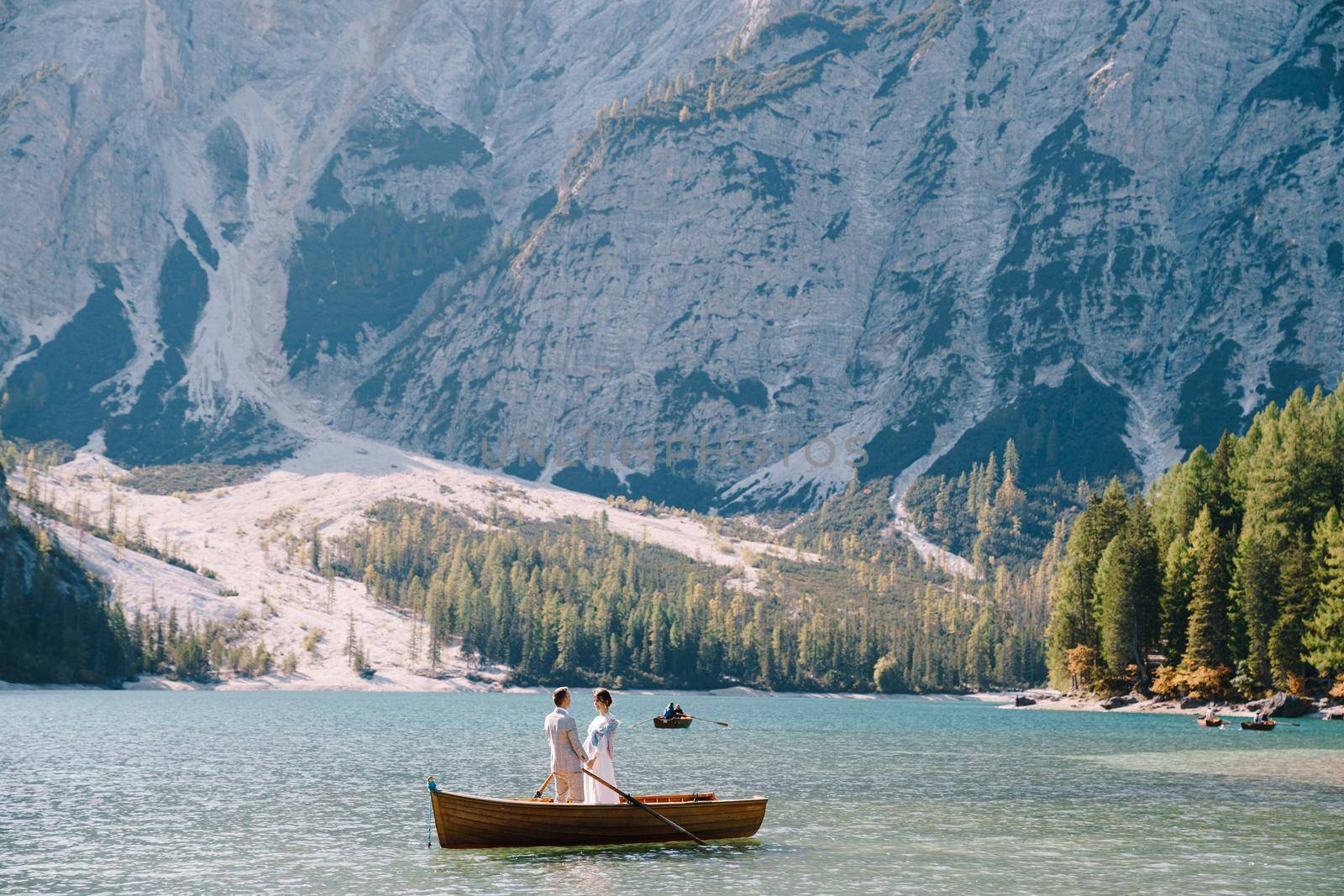 The bride and groom are sailing in a wooden boat with oars at the Lago di Braies in Italy. Wedding in Europe, on Braies lake. Newlyweds are standing embracing in a boat. by Nadtochiy