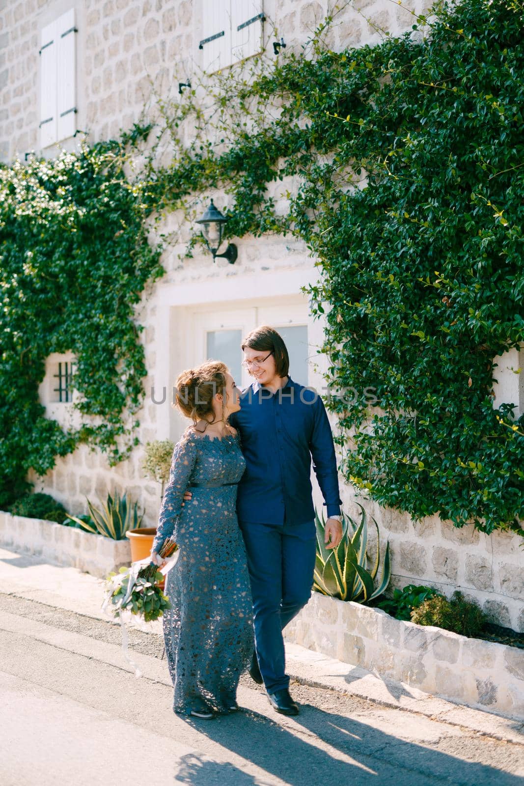 The bride and groom are embracing along the road near an old building with a white door, the wall is entwined with a green liana . High quality photo