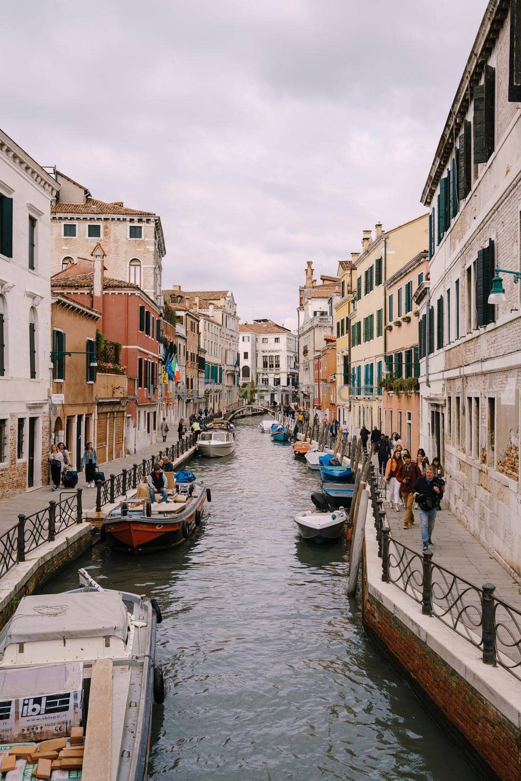 A classic view of the narrow sea Venetian canal. Colorful buildings stand opposite each other, boats moored near the shore, gondolier rides tourists on a gondola. Blue skies on a sunny day.