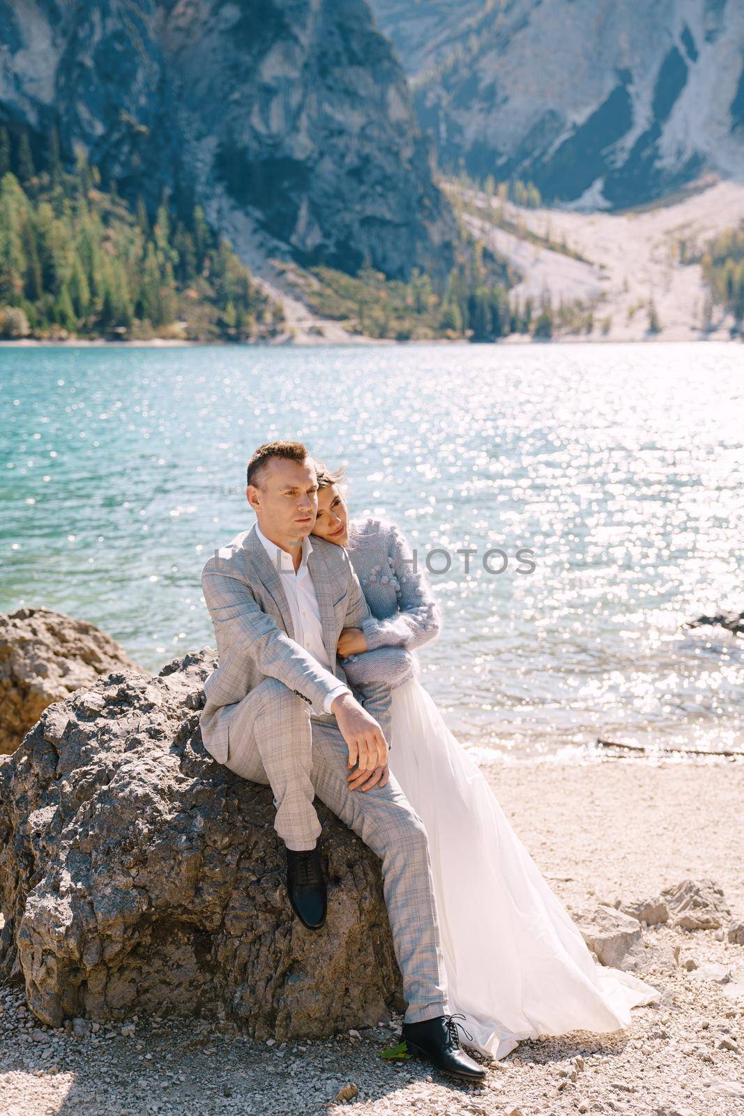 Bride and groom sit embracing against background of stones at the Lago di Braies in Italy. Destination wedding in Europe, on Braies lake. Loving newlyweds walk against the backdrop of amazing nature by Nadtochiy