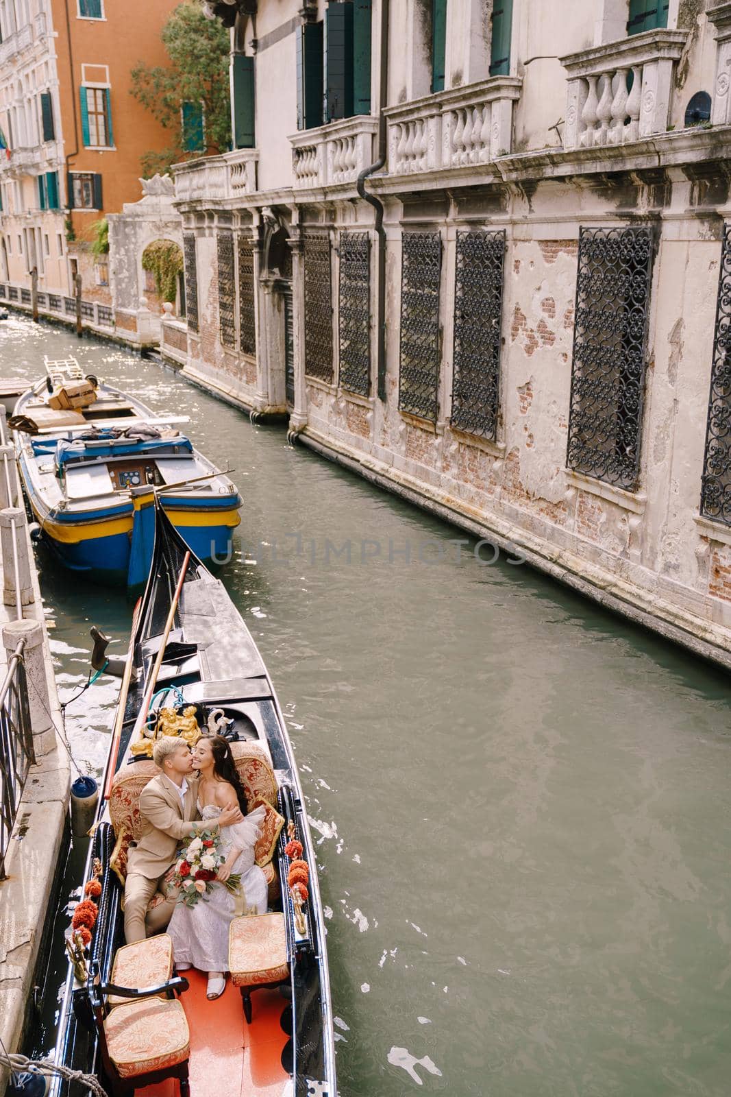 Italy wedding in Venice. A gondolier rolls a bride and groom in a classic wooden gondola along a narrow Venetian canal. Newlyweds kiss in a boat moored to the shore. by Nadtochiy