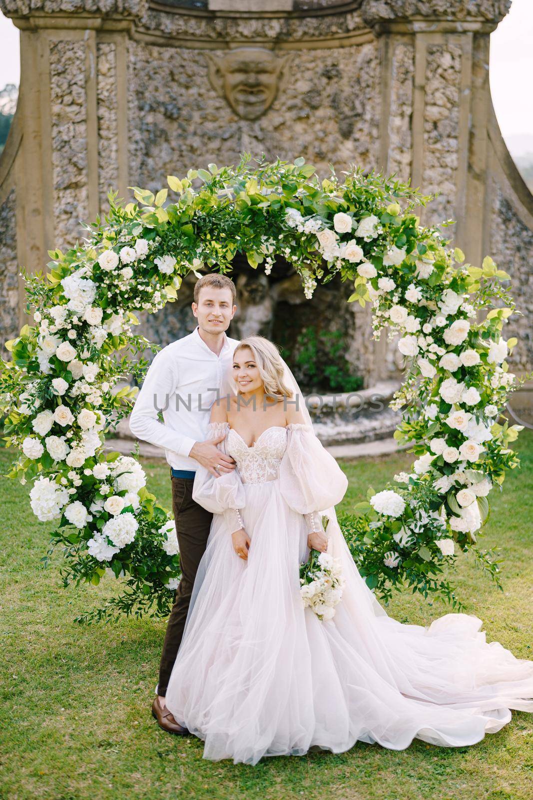 Wedding couple near round wedding arch decorated with white flowers and greenery in front of an ancient Italian architecture. Wedding at an old winery villa in Tuscany, Italy