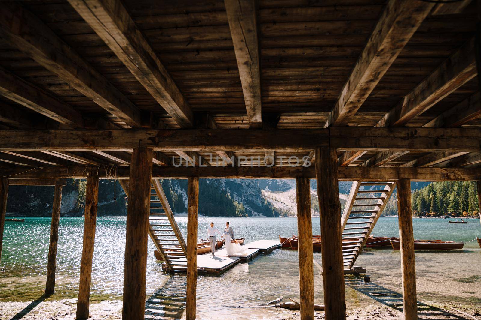 The bride and groom walk along a wooden boat dock at the Lago di Braies in Italy. Wedding in Europe, on Braies lake. Newlyweds walk, kiss, hug on a background of rocky mountains. by Nadtochiy