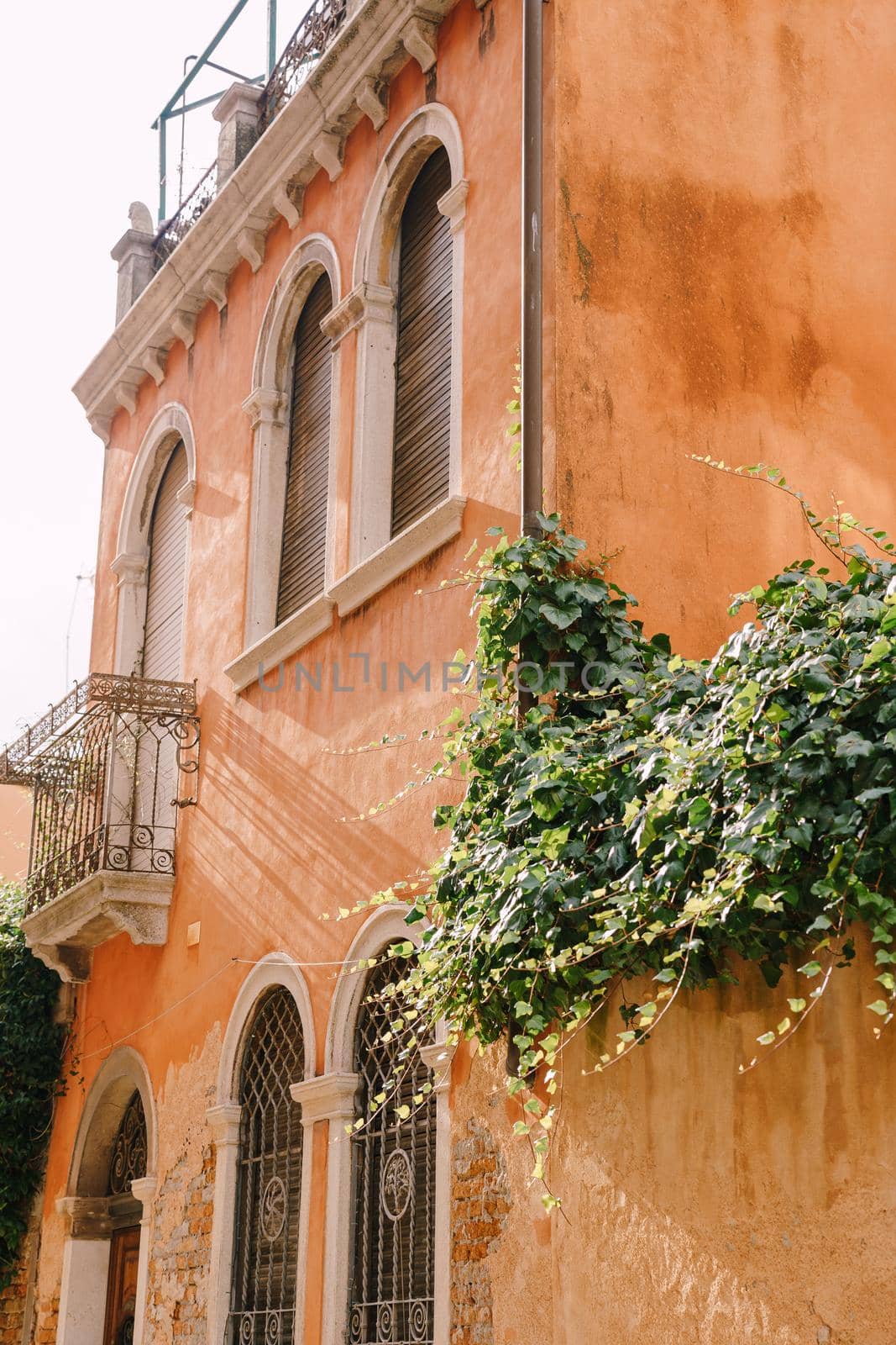 An ancient building in Venice, Italy, close-up. Peach-colored walls, classic Venetian arched windows with wooden closed rolls, a small balcony with a metal forged grille.