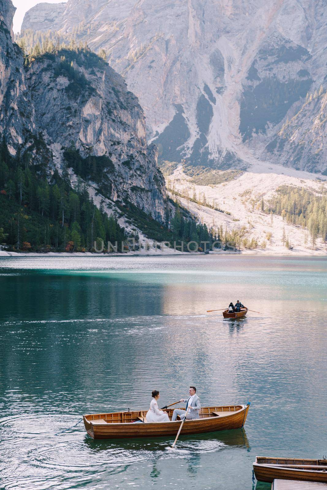 Wedding couple swim in a wooden boat on Lago di Braies in Italy. Newlyweds in Europe, at Braies Lake, in the Dolomites. The groom rows with oars, the bride sits opposite him.