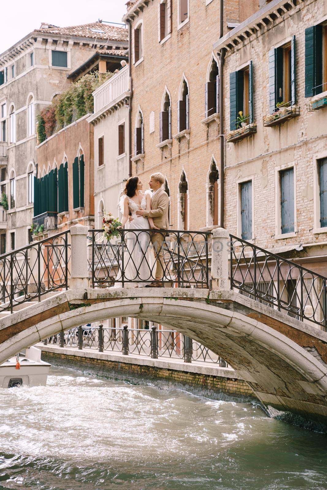 Italy wedding in Venice. The bride and groom are standing on a stone bridge over a narrow Venetian canal. Newlyweds walk around the city taking pictures on the street. by Nadtochiy
