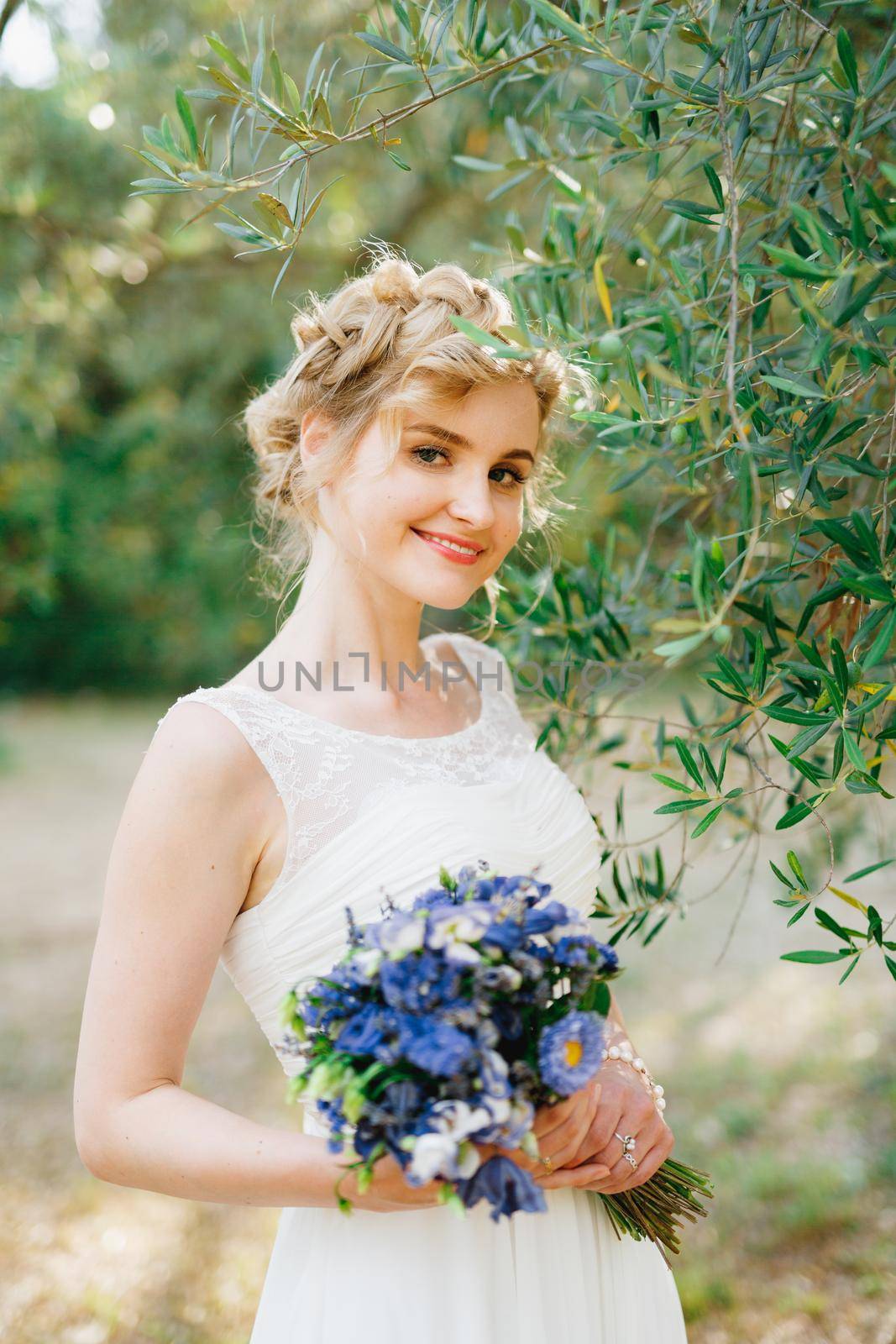 A tender bride with a bouquet of blue flowers in her hands stands by green olive branches in a grove and smiles by Nadtochiy
