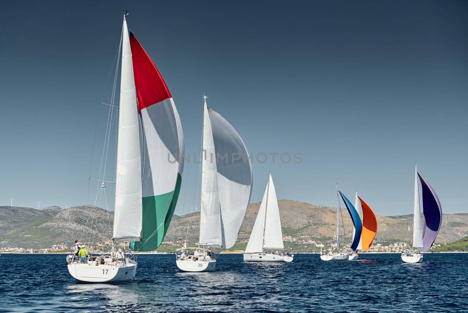 Sailboats compete in a sail regatta at sunset, race of sailboats, reflection of sails on water, multicolored spinnakers, number of boat is on aft boats, island is on background, clear weather by vladimirdrozdin