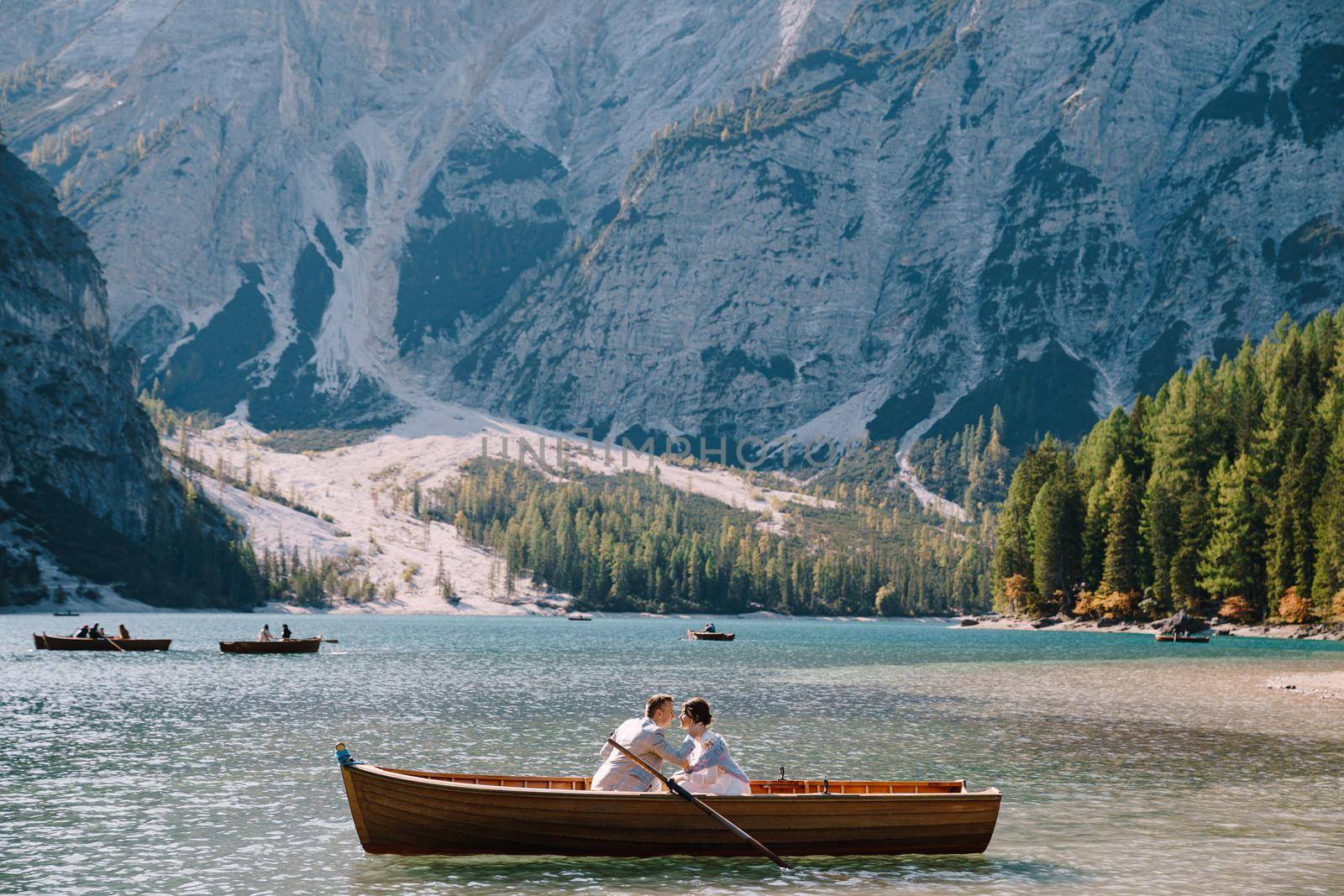 Bride and groom sail in a wooden boat at the Lago di Braies in Italy. Wedding in Europe, on Braies lake. The newlyweds are sitting in the boat and kissing. by Nadtochiy