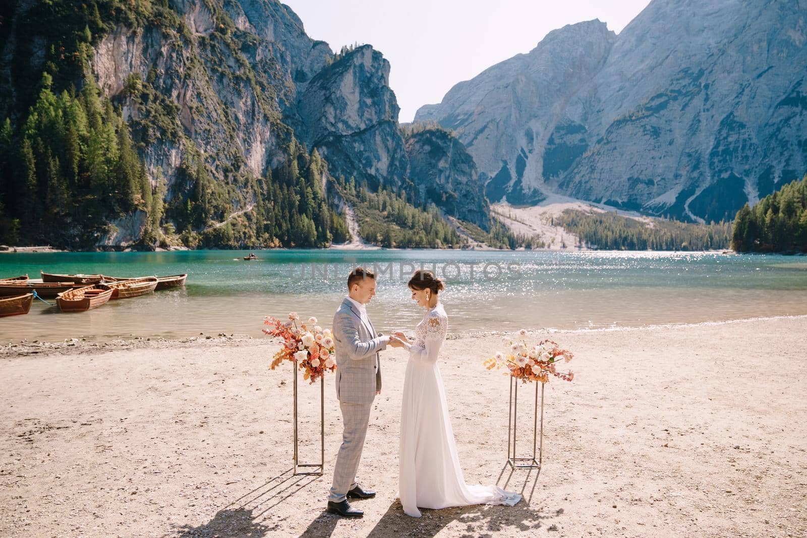 The bride dons a ring to the groom at the venue for the ceremony, with an arch of autumn floral columns, against the backdrop of Lago di Braies in Italy. Destination wedding in Europe, at Braies lake.
