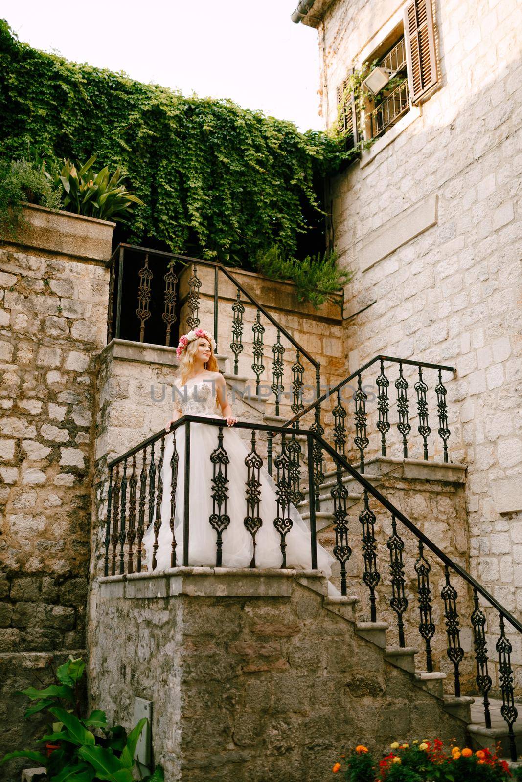 A bride with a wreath of white and pink roses stands on the staircase with a wrought railing in the old town of Kotor by Nadtochiy