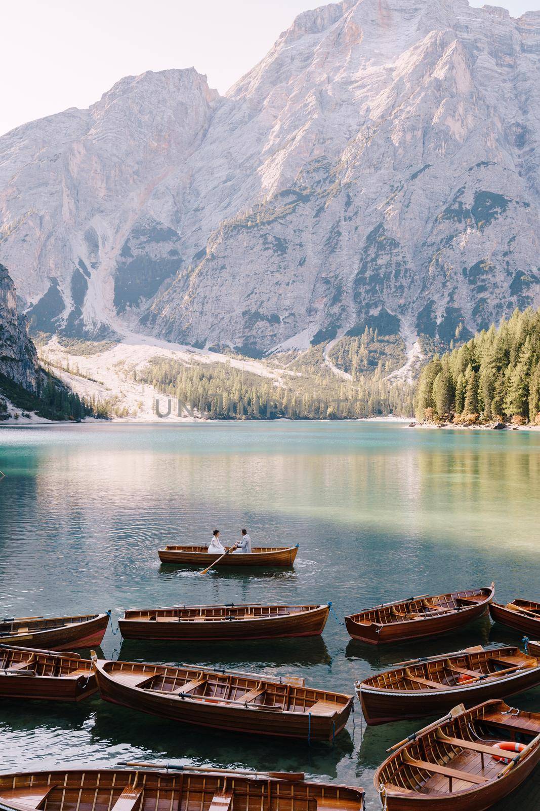 Wedding couple sailing in a wooden boat at the Lago di Braies in Italy. Newlyweds in Europe, on Braies lake, in the Dolomites. The groom rows the oars, the bride sits opposite him. by Nadtochiy