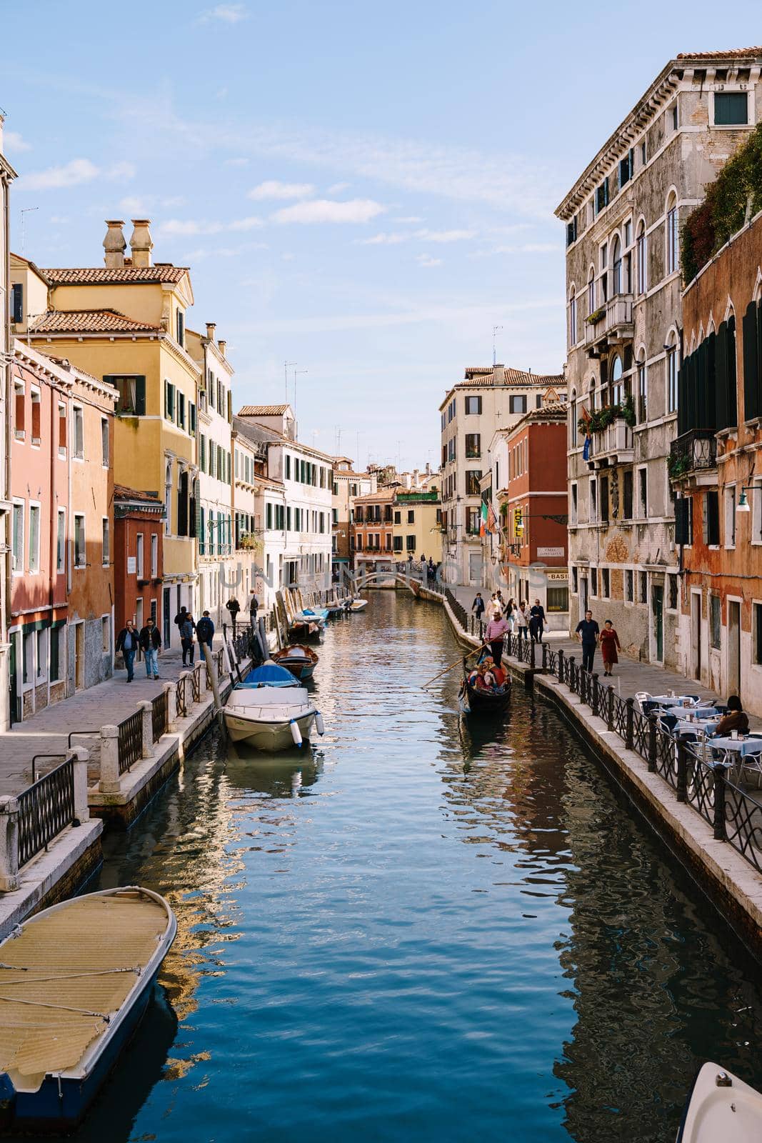 A classic view of the narrow sea Venetian canal. Colorful buildings stand opposite each other, boats moored near the shore, gondolier rides tourists on a gondola. Blue skies on a sunny day.