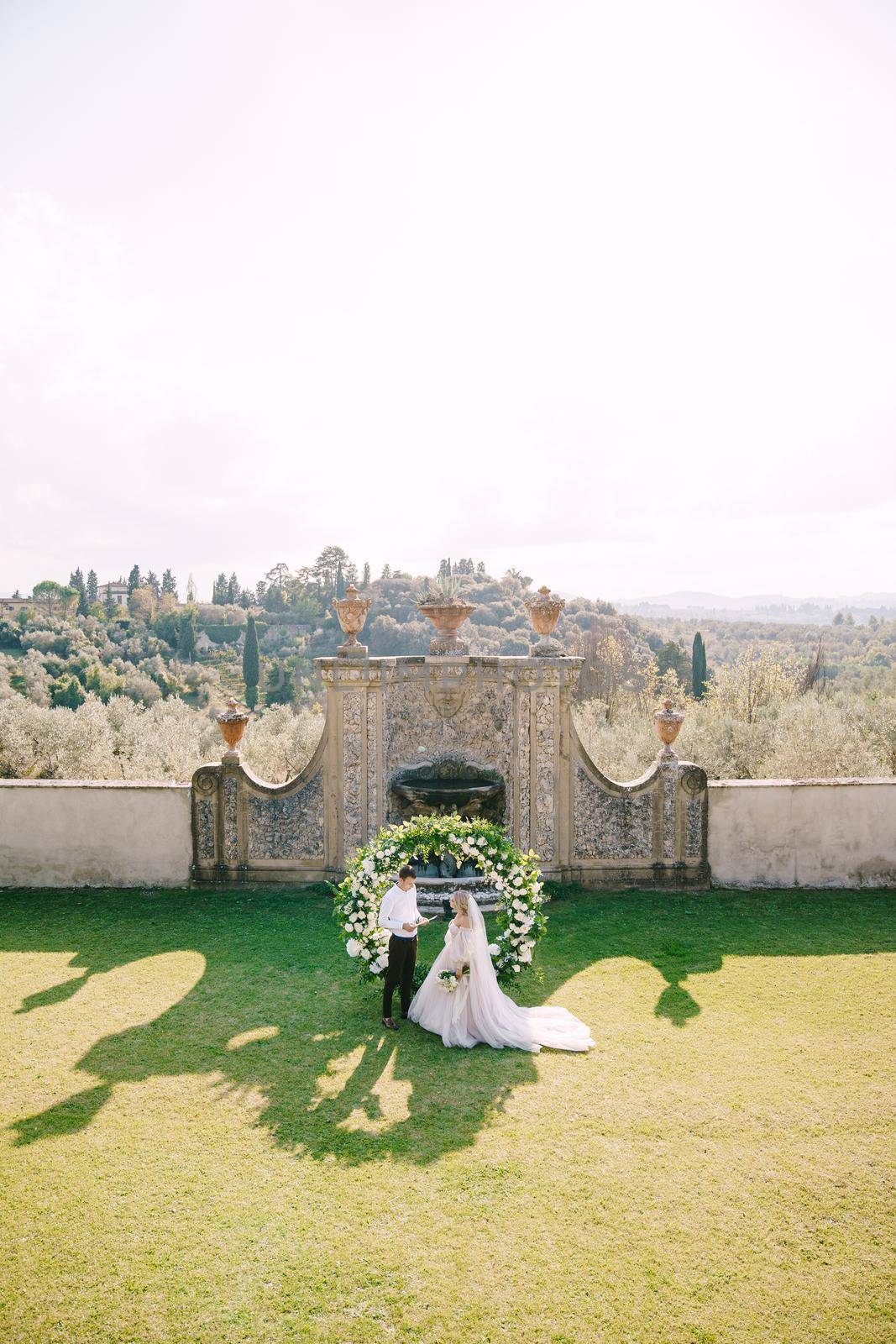Florence, Italy - 01 october 2019: Wedding at an old winery villa in Tuscany, Italy. Wedding couple under a round arch of flowers. The groom reads wedding vows. by Nadtochiy