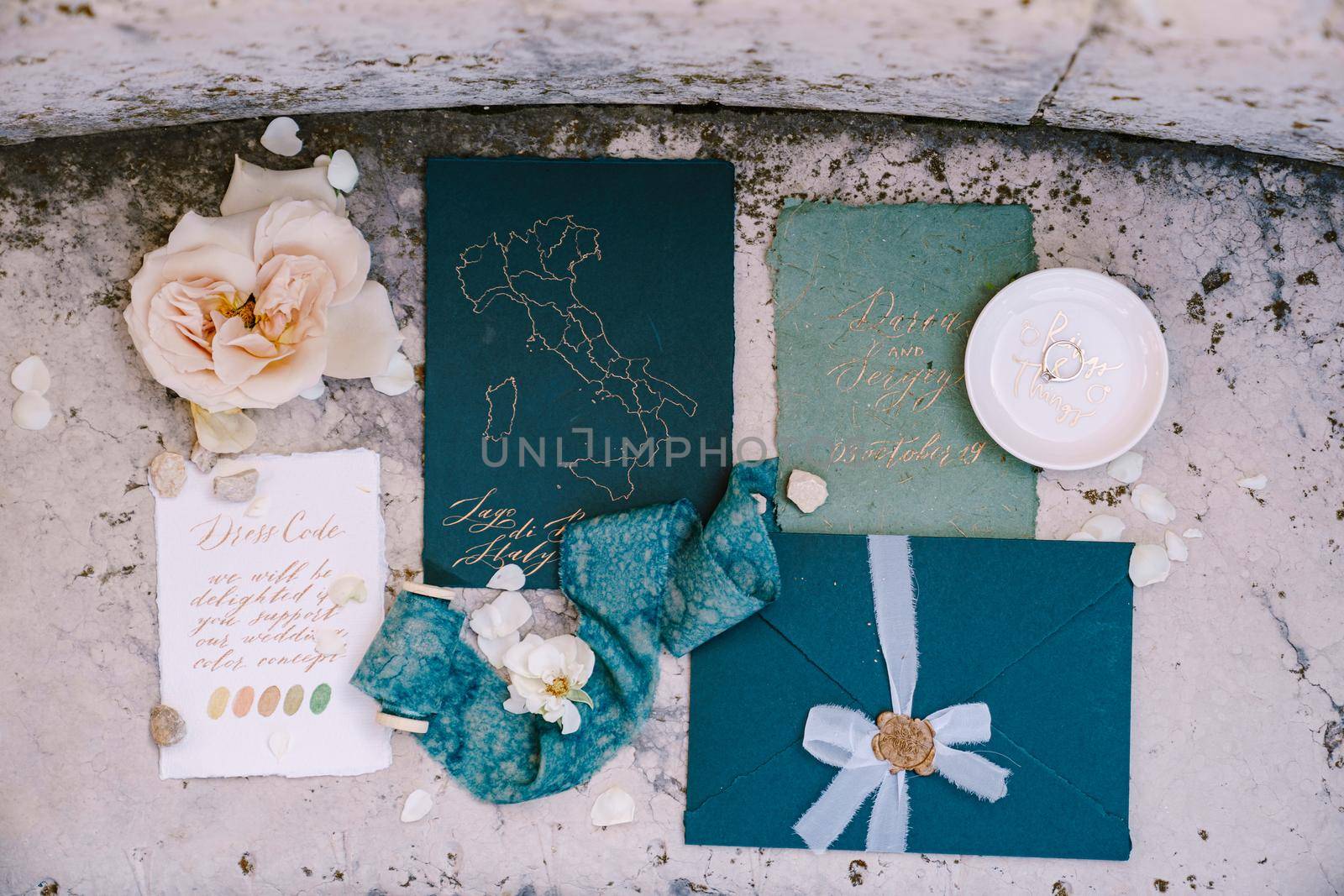 Blue mockup of invitations to wedding, blue envelope with seal print of sealing wax, a silk blue ribbon, a white sheet with the colors of dess code, pink flower against background of marble stone