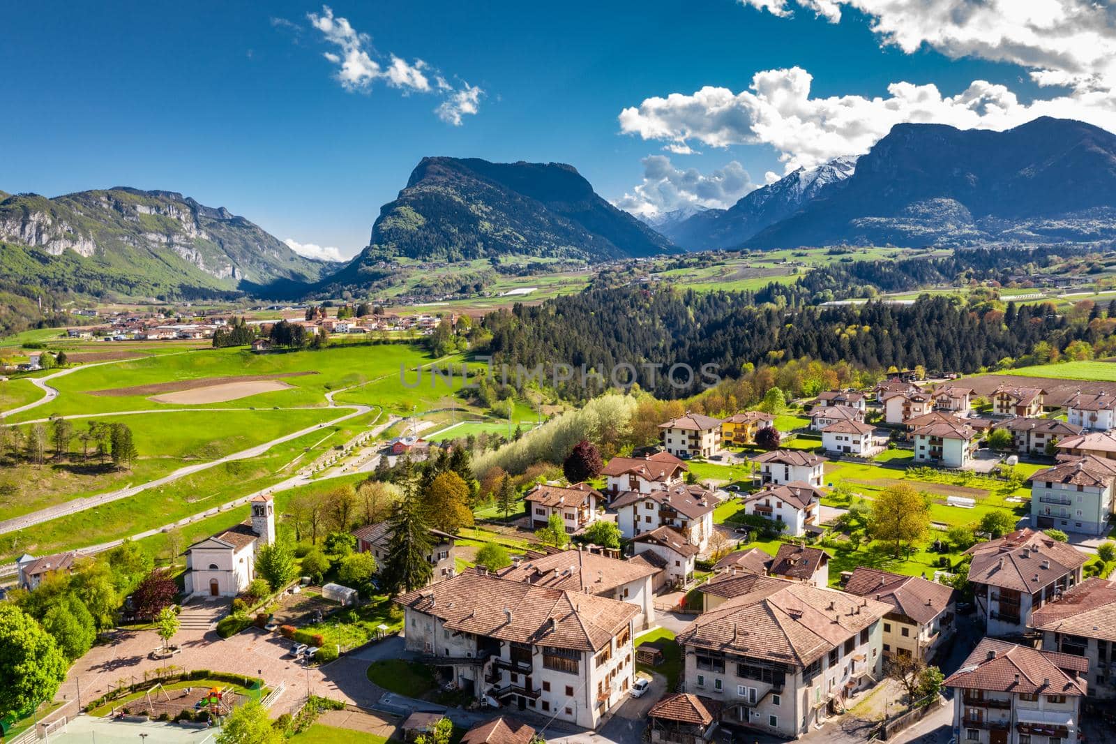 Aerial view of improbable green meadows of Italian Alps, Comano Terme, huge clouds over a valley, roof tops of houses, Dolomites on background, sunshines through clouds by vladimirdrozdin