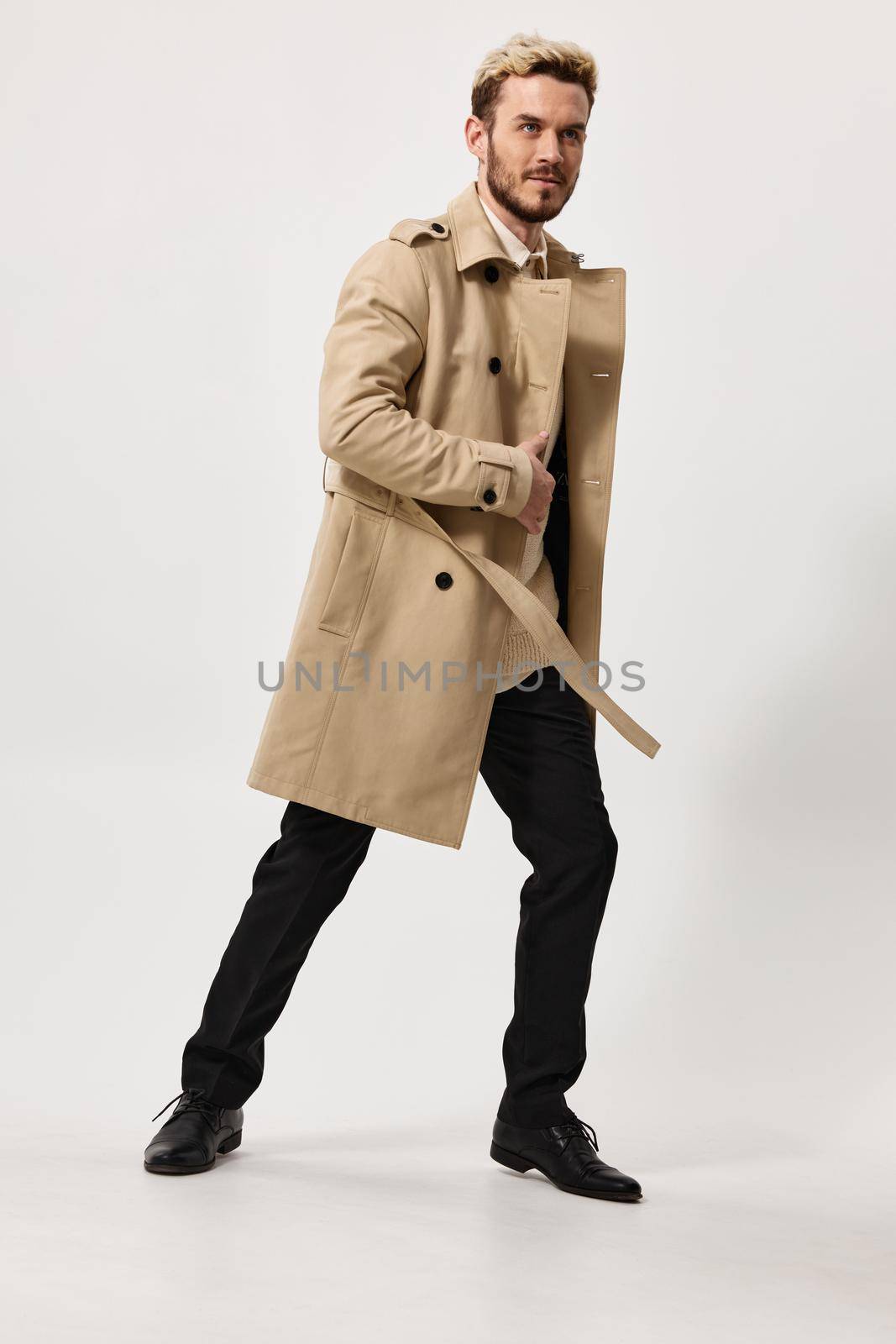 an energetic man in trousers and a beige coat leaned to the side against a light background in full growth. High quality photo