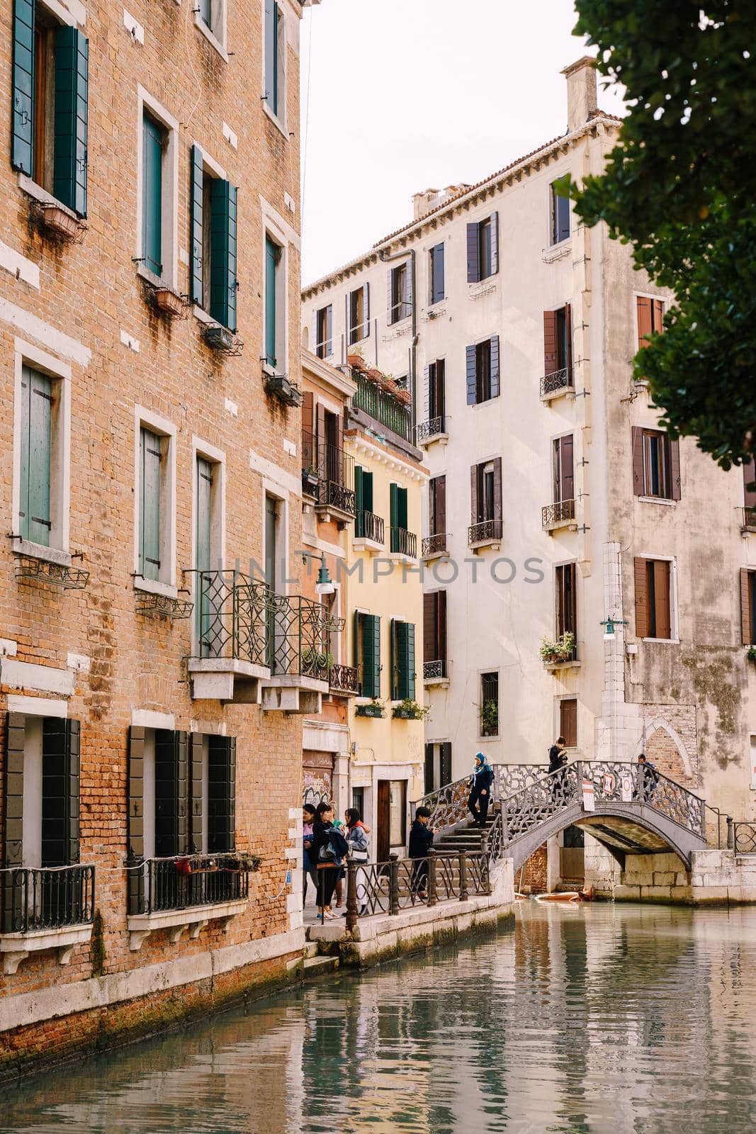 Tourists walk on an arable bridge over a small Venetian canal, against the backdrop of apartment buildings on streets of Venice, Italy.