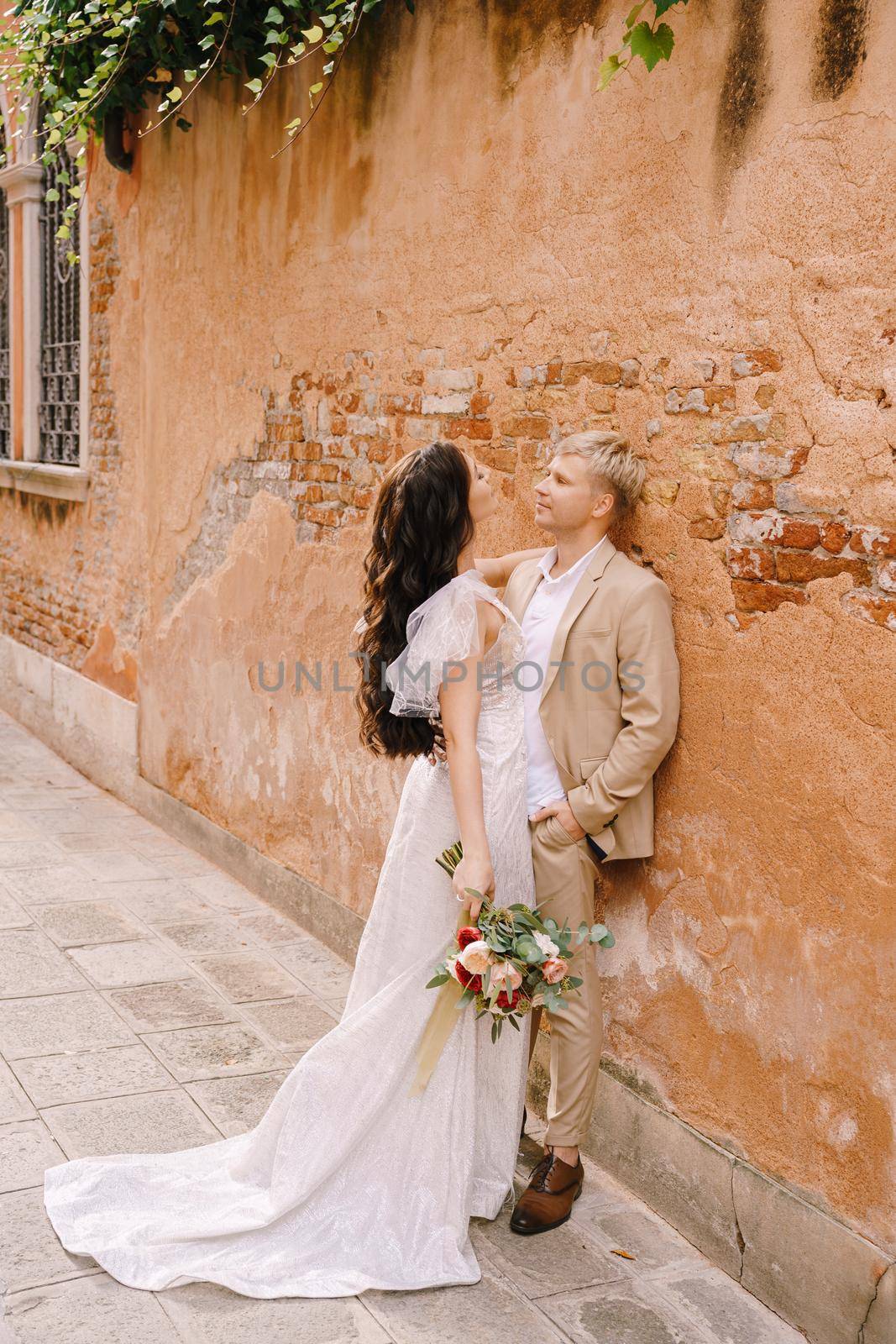 The bride and groom walk through the deserted streets of the city. Newlyweds stand next to each other and cuddle near the orange wall of a beautiful house.