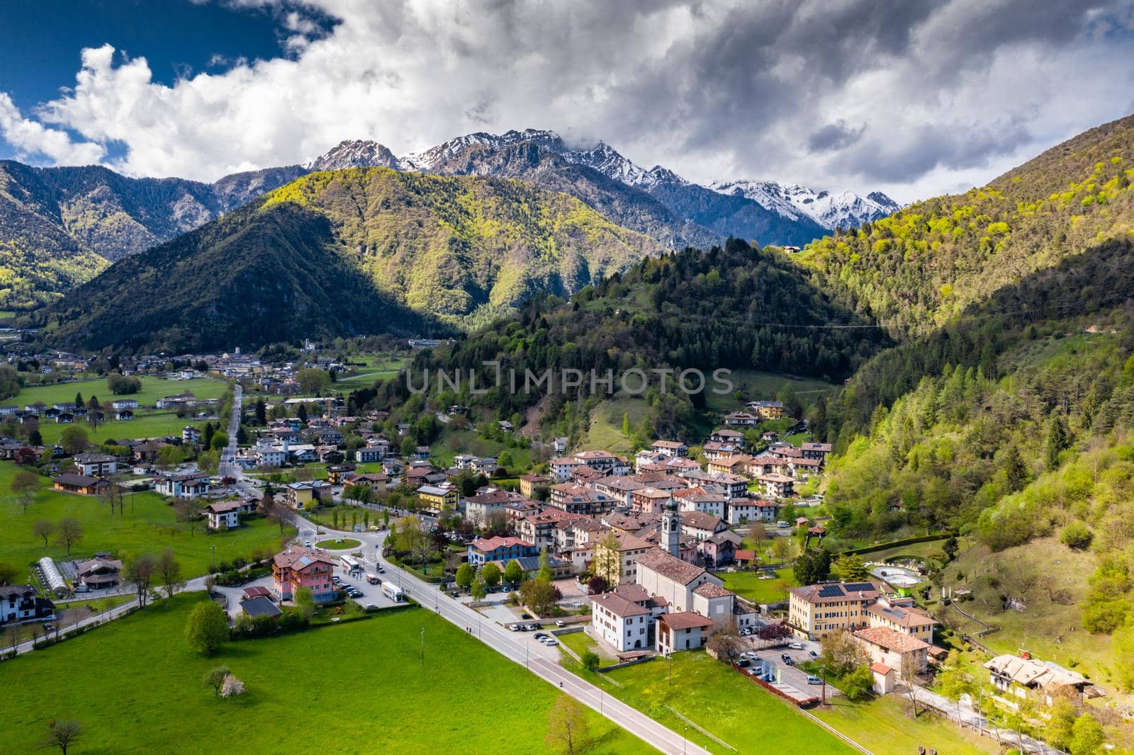 Aerial view of improbable green meadows of Italian Alps, Comano Terme, huge clouds over a valley, roof tops of houses, Dolomites on background, sunshines through clouds by vladimirdrozdin