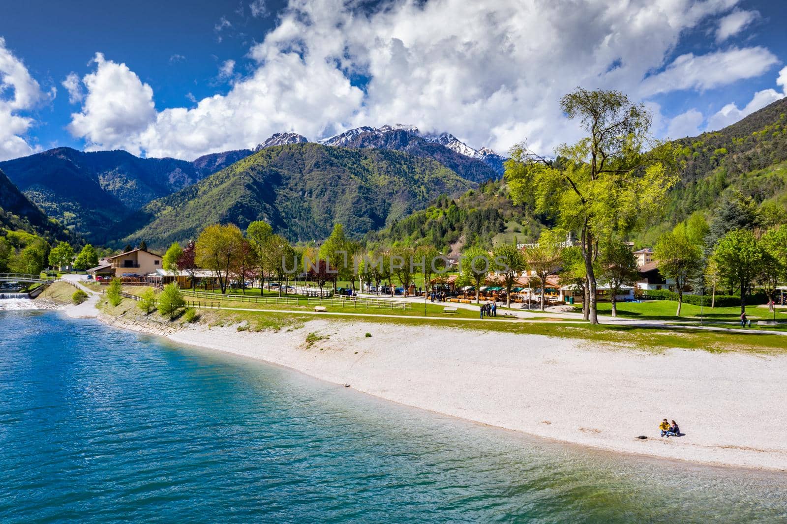 The Improbable aerial landscape of village Molveno, Italy, azure water of lake, The couple sits on a beach, snow covered mountains Dolomites on background, sunny weather, a piers, coastline by vladimirdrozdin