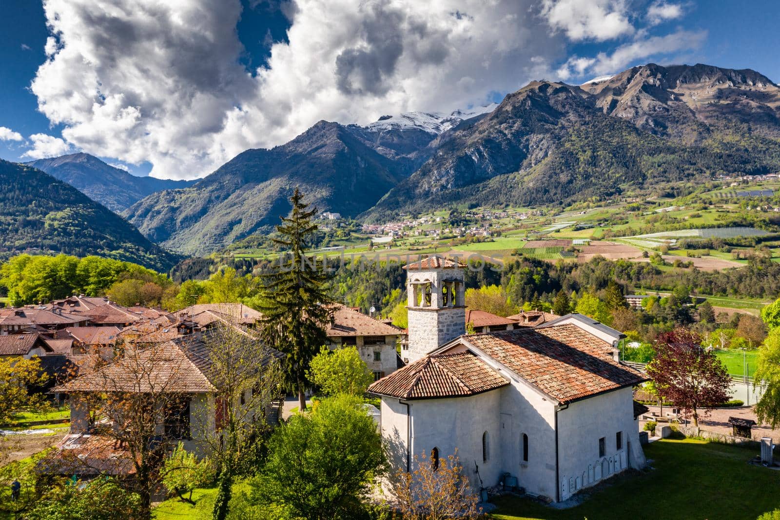 Aerial view of valley with church in Cares, Trentino, green slopes of the mountains of Italy, huge clouds over a valley, Dolomites on background