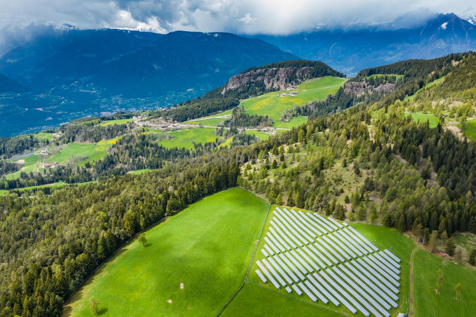 Aerial view of farm of solar batteries on green slopes of the mountains of Italy, Trentino, huge clouds over a valley, roofs of houses of settlements, green meadows, a clear energy, energy of the sun, drone view point