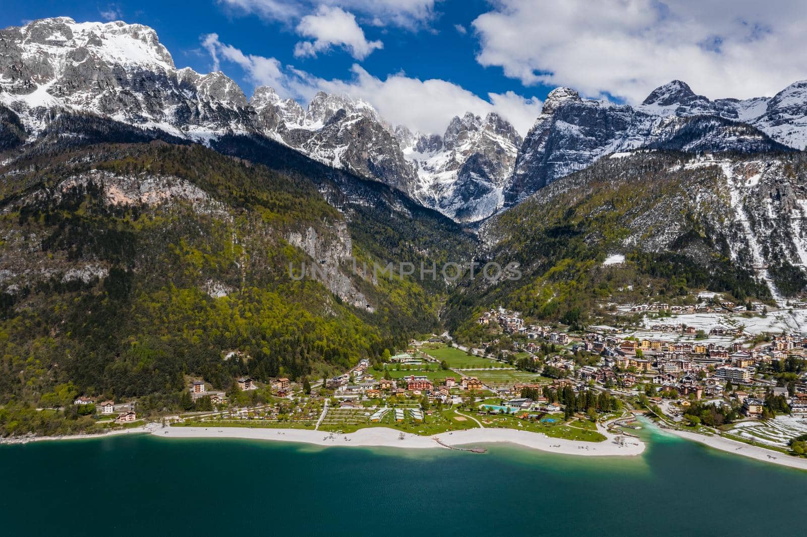 The Improbable aerial landscape of village Molveno, Italy, azure water of lake, empty beach, snow covered mountains Dolomites on background, roof top of chalet, sunny weather, a piers, coastline, slopes