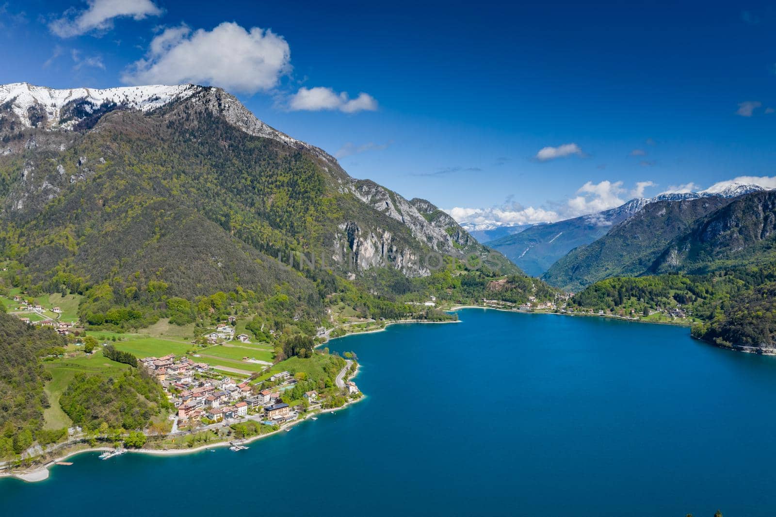 The Improbable aerial landscape of village Molveno, Italy, azure water of lake, empty beach, snow covered mountains Dolomites on background, roof top of chalet, sunny weather, a piers, coastline, slopes