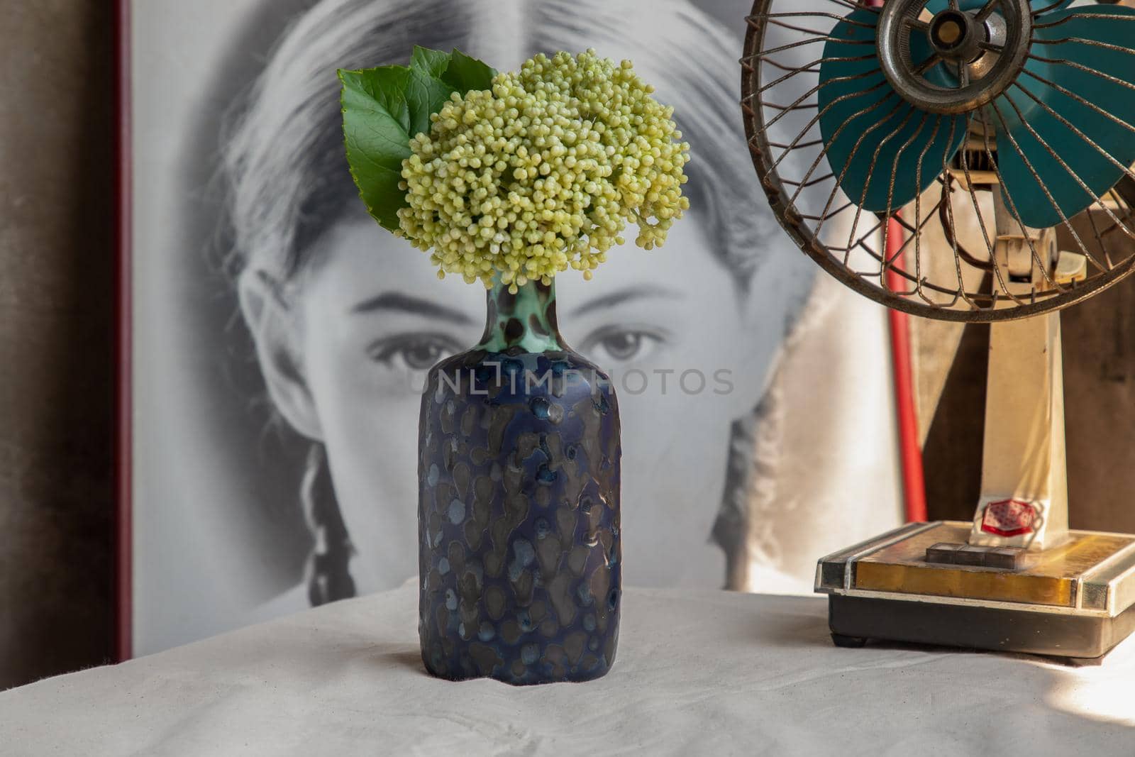 Bangkok, Thailand -Feb 06, 2021 : Bouquet of flowers in Blue ceramic vase and vintage fan on white textured table cloth in front of Classic chinese poster movie frame with old cement wall. Home decor, Selective focus.