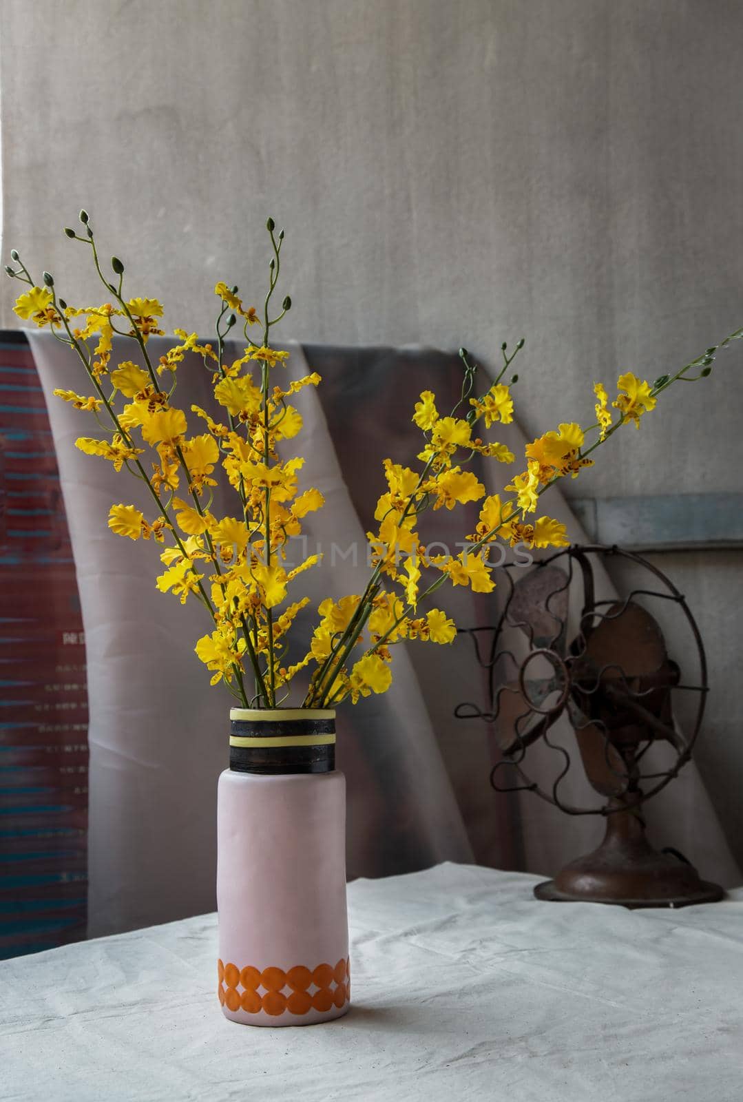 Bouquet of yellow flowers in Handmade ceramic vase and Vintage fan on white textured table cloth with old cement wall. Home decor, No focus, specifically.