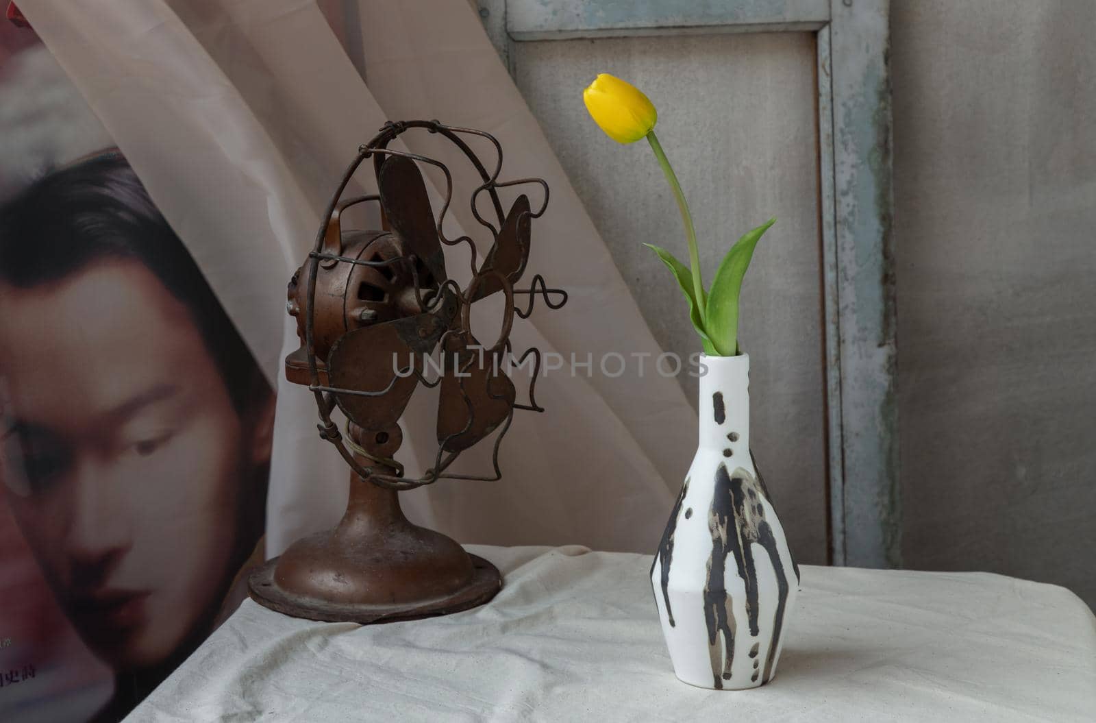 Bangkok, Thailand - Feb 06, 2021 : Yellow flower in Black and White Handmade ceramic vase and Vintage fan on white textured table cloth in front of Classic chinese poster movie frame with old cement wall. Home decor, Selective focus.